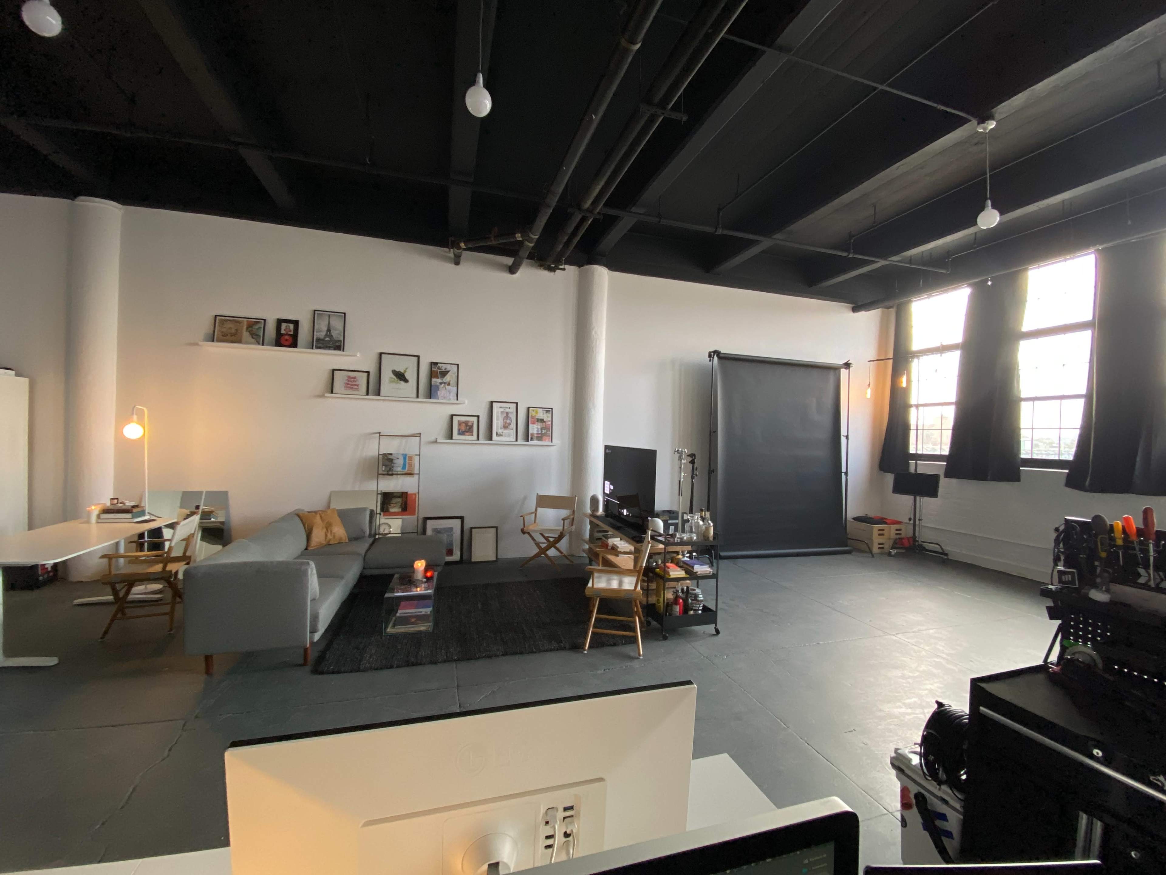 A spacious indoor studio with a gray sofa, a workspace, a large screen setup, and large windows letting in natural light.