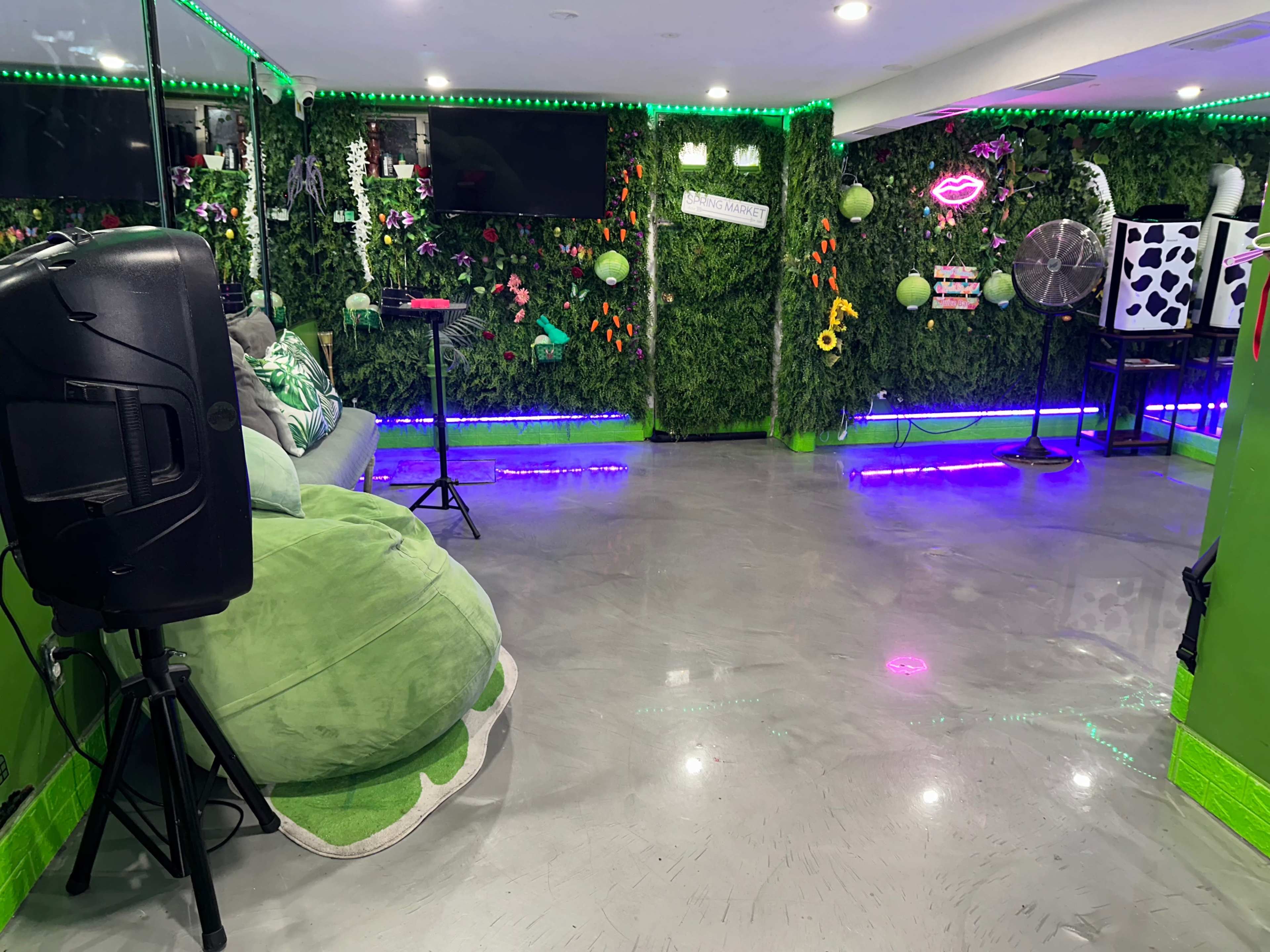 The room features green walls adorned with artificial plants, neon lights, and a variety of decorative elements, including a speaker and a fan.