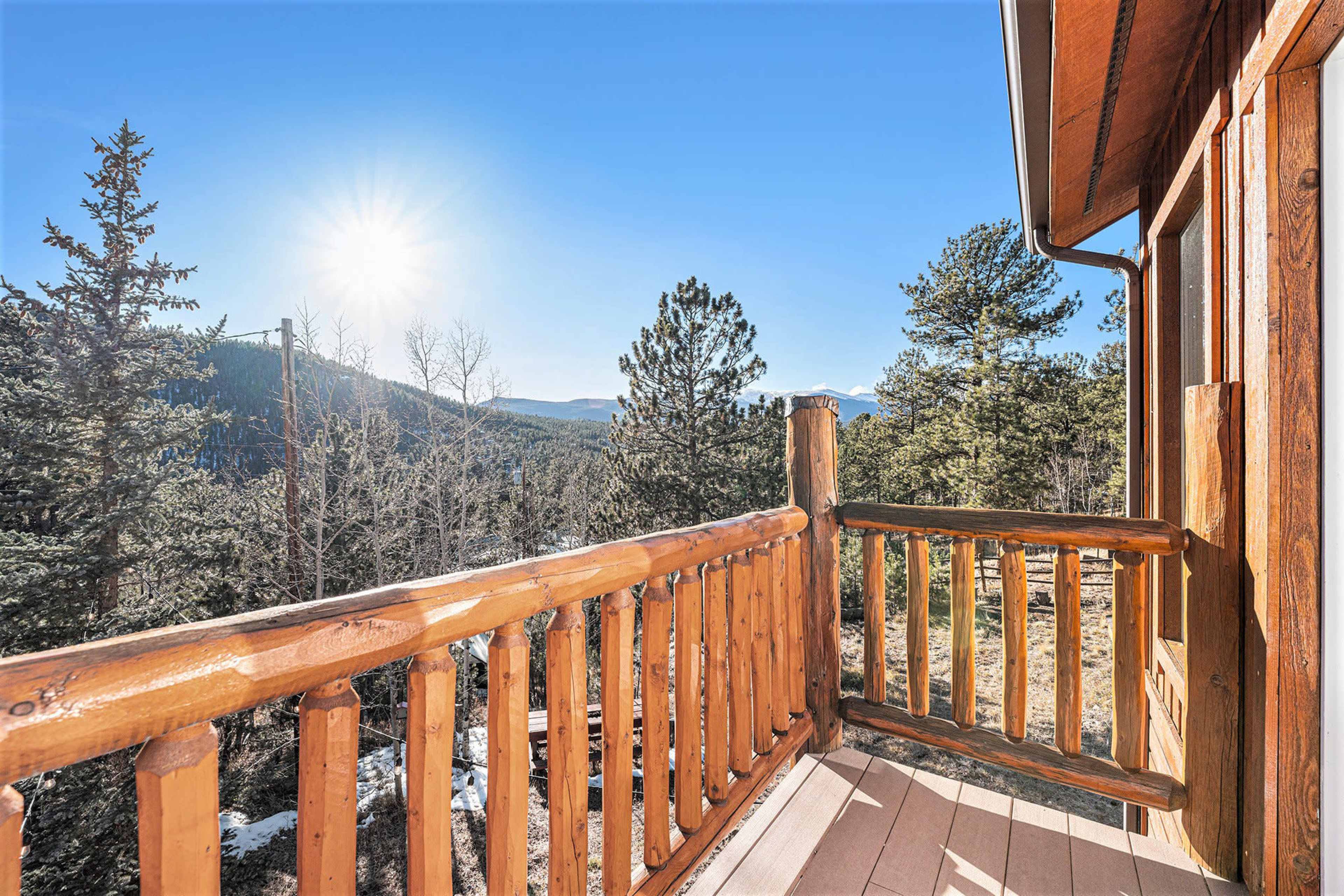 The image shows a wooden balcony with a railing overlooking a mountainous landscape under a clear blue sky with the sun shining.