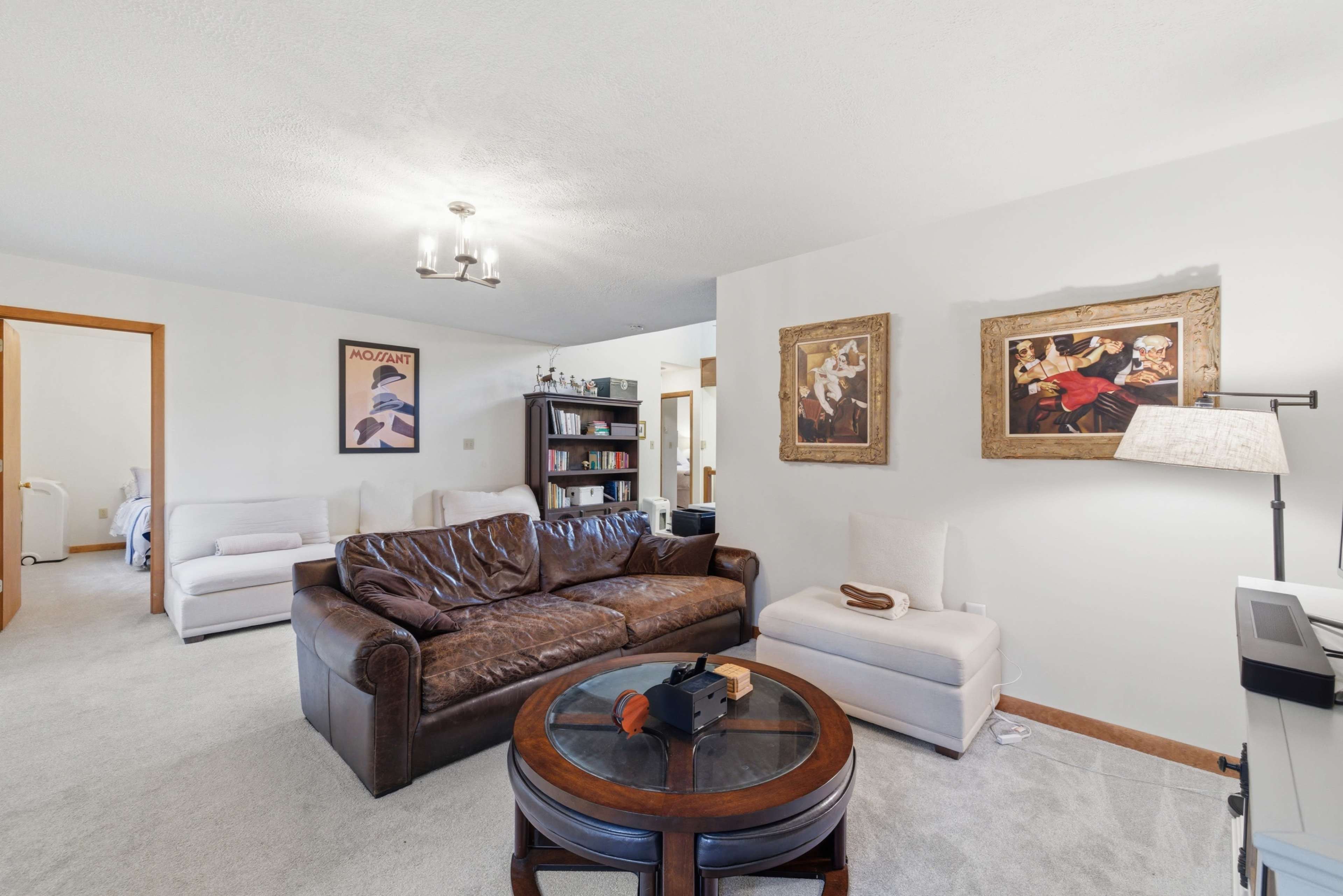 The image shows a living room furnished with a brown leather sofa, two white chairs, a circular wooden table, and framed artwork on the walls.