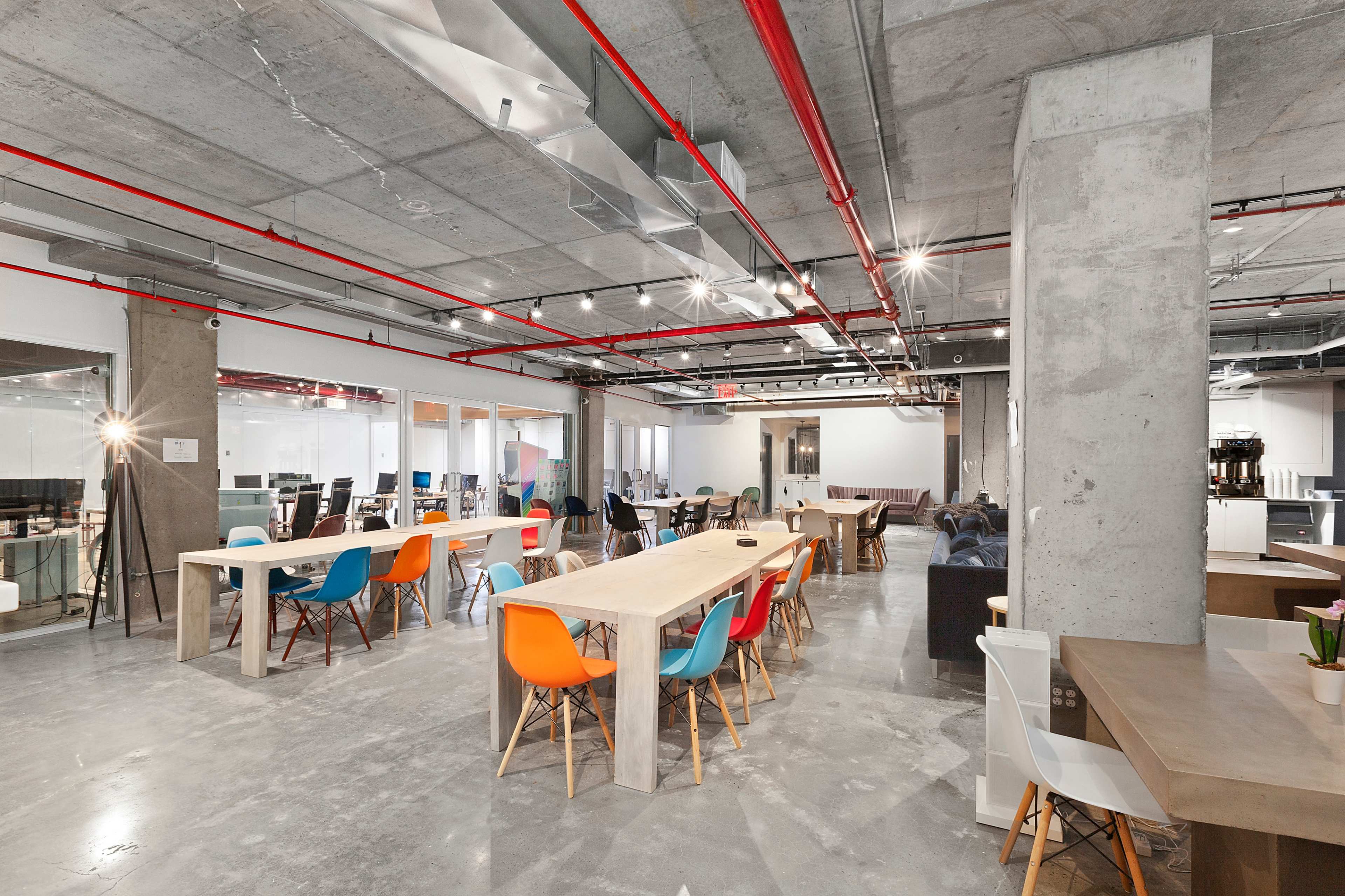 The image shows a modern, open office space with long tables, colorful chairs, exposed concrete walls, and visible overhead piping.
