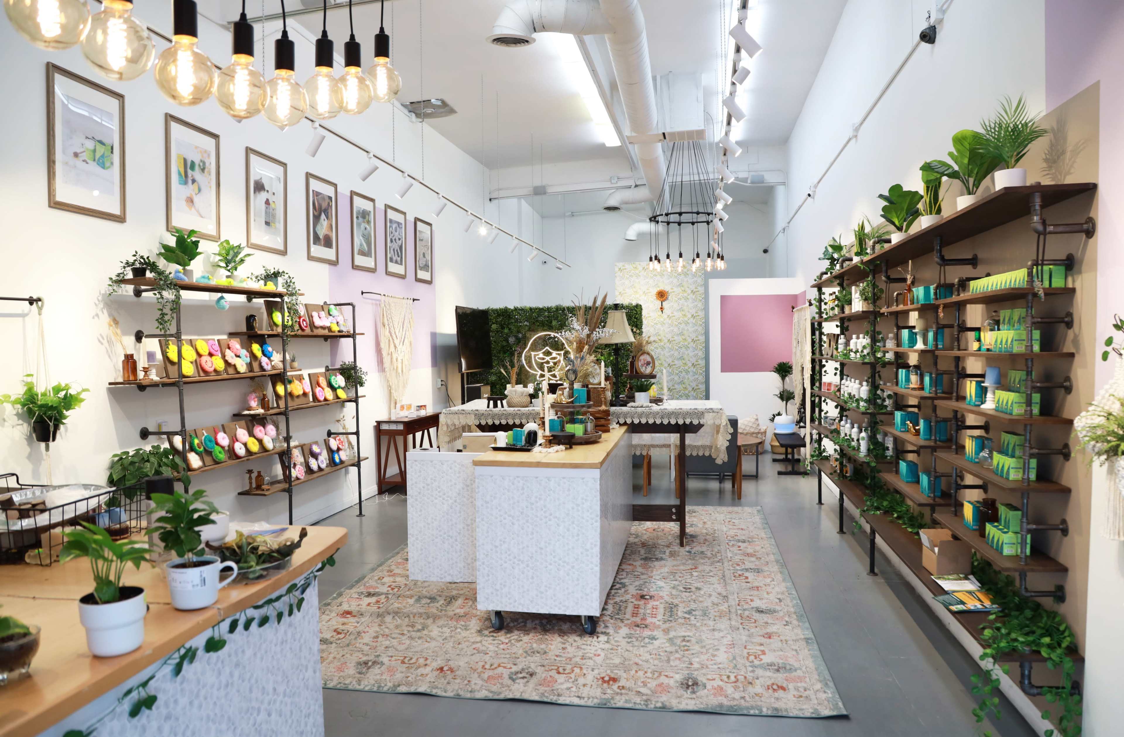A brightly lit retail space with shelves filled with various colorful products, a central table with decorative arrangements, and plants throughout the room.