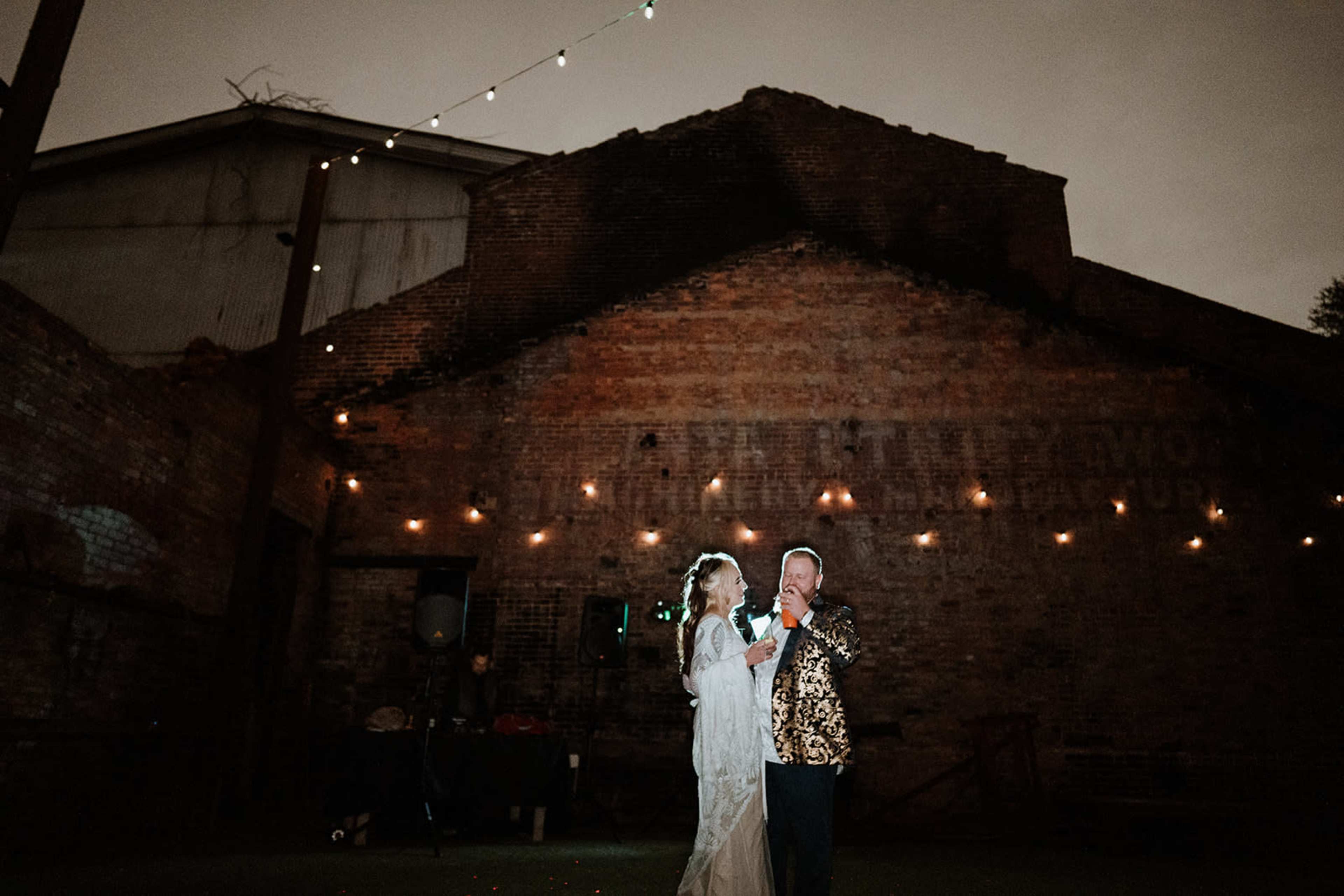 A couple stands together on an outdoor stage illuminated by soft lights, with a rustic brick wall and string lights in the background.