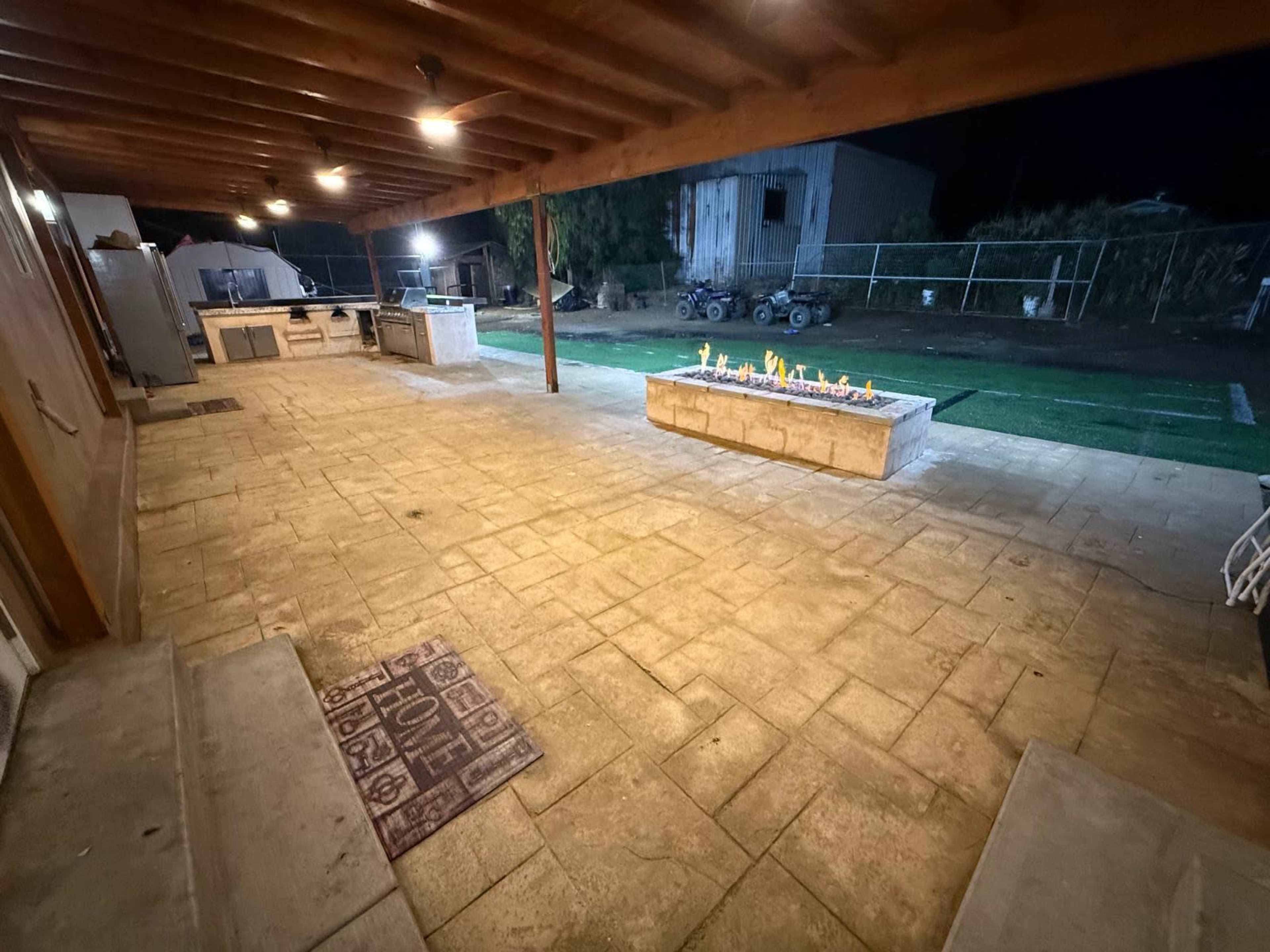 The image shows a spacious outdoor patio area at night, featuring a barbecue setup, a stone fire pit with flames, and a dirt yard in the background.