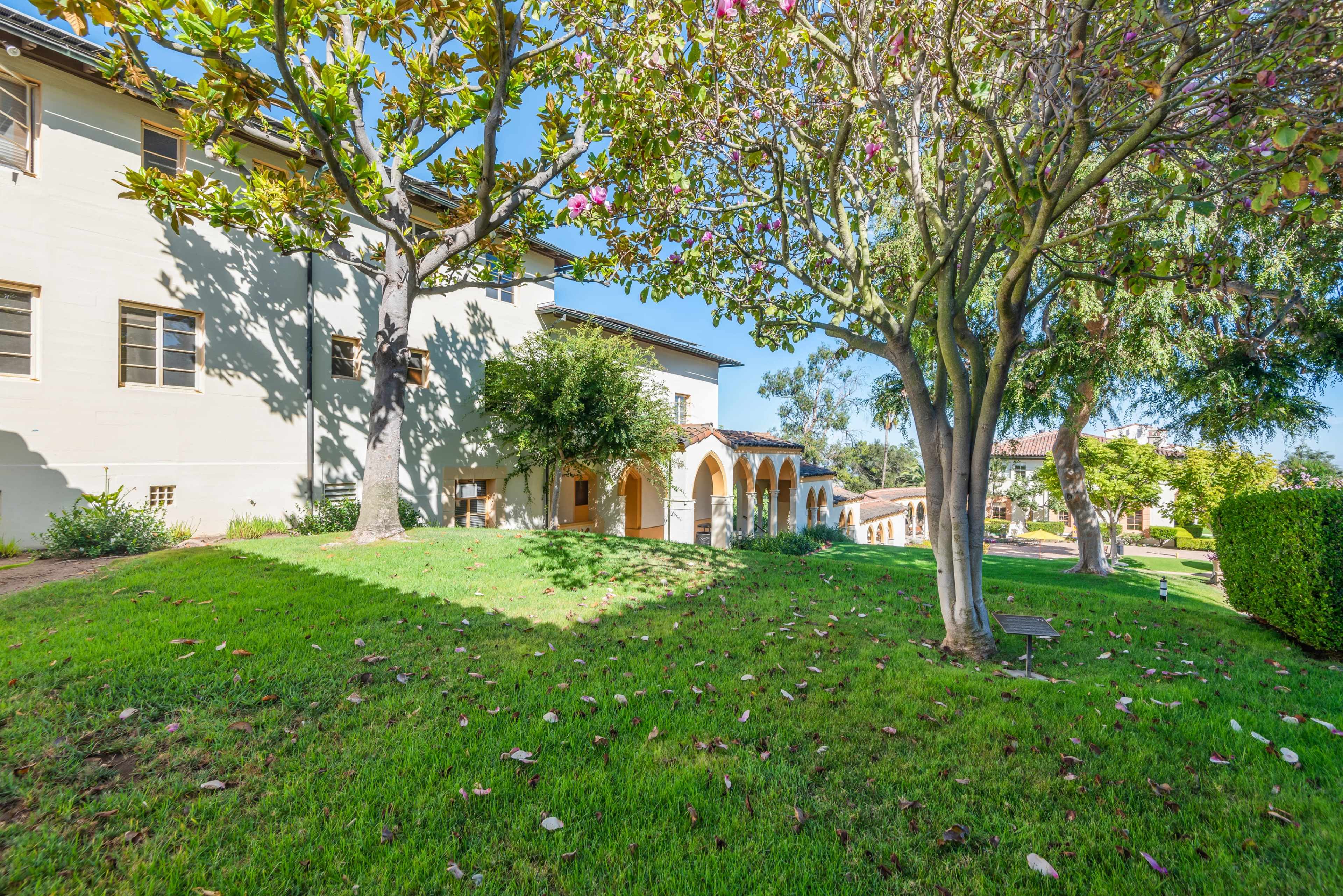 The image shows a grassy area surrounded by trees and a two-story building with arched doorways.