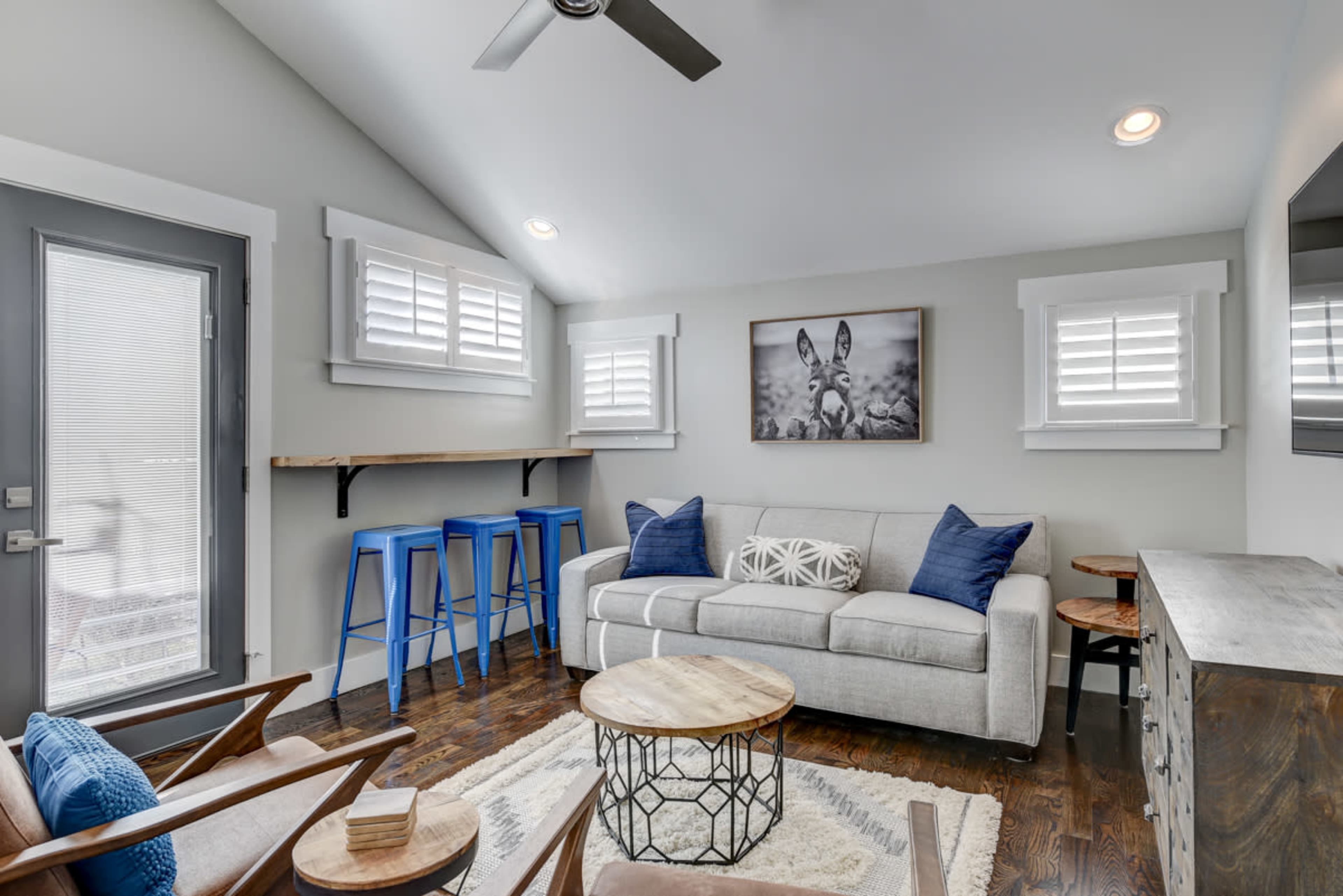 A modern living room with a gray sofa, wooden coffee table, two armchairs, and a wall-mounted television, alongside a set of blue bar stools near a window.