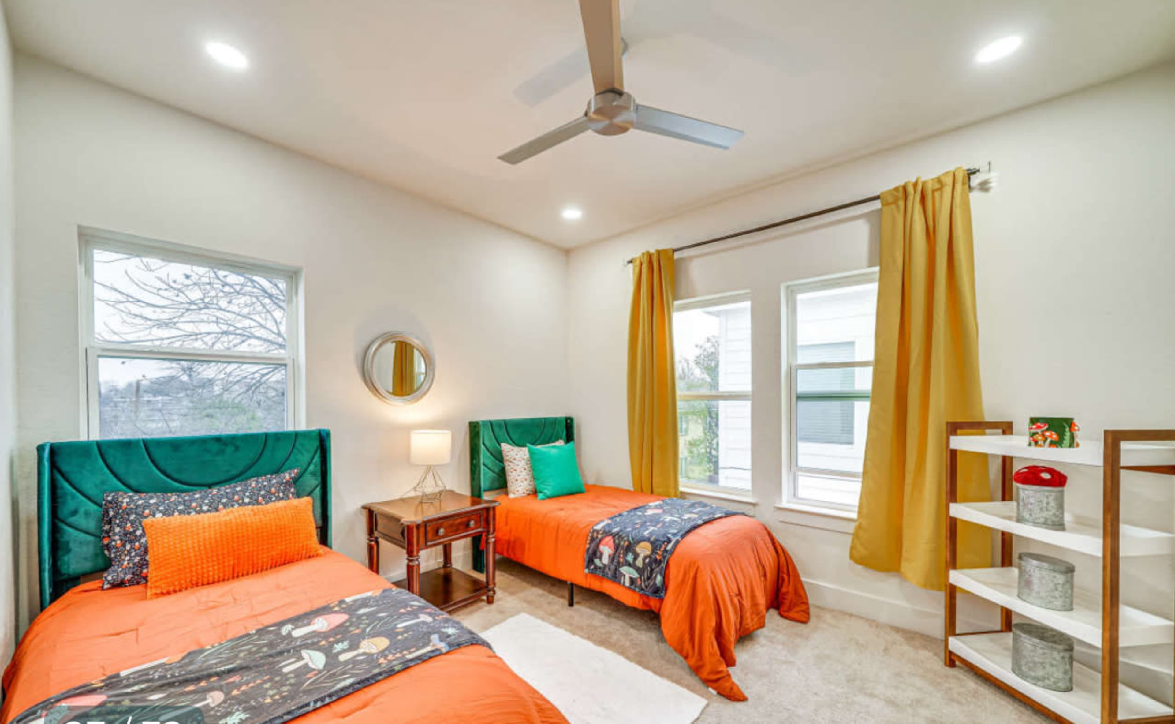 The image shows a brightly lit bedroom featuring two twin beds with orange bedding and a nightstand between them, along with a shelving unit and large window dressed with yellow curtains.