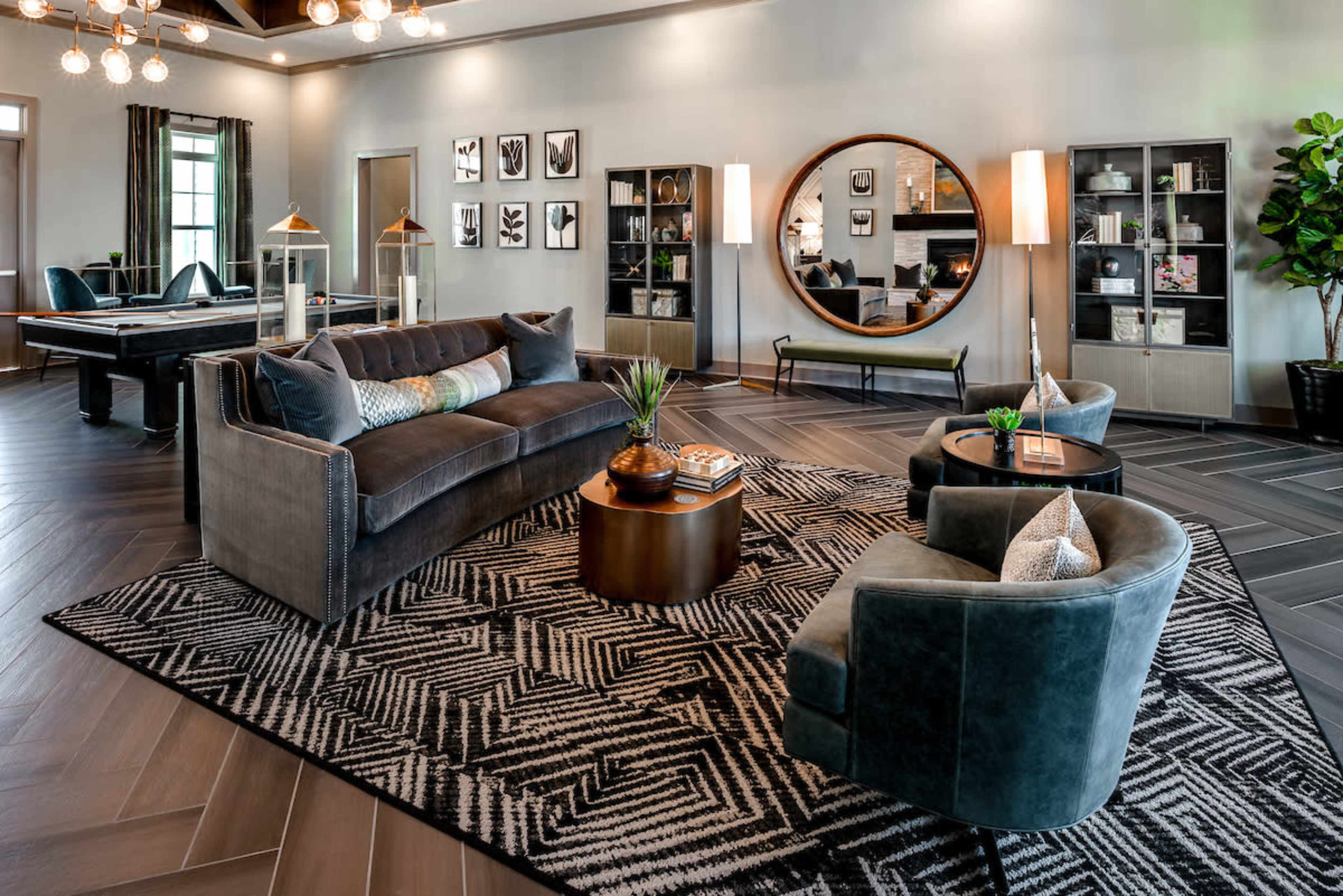 The image shows a modern interior living space featuring a gray sofa, two round chairs, a circular mirror, and various decorative elements on shelves, all set on a patterned rug with dark wooden flooring.