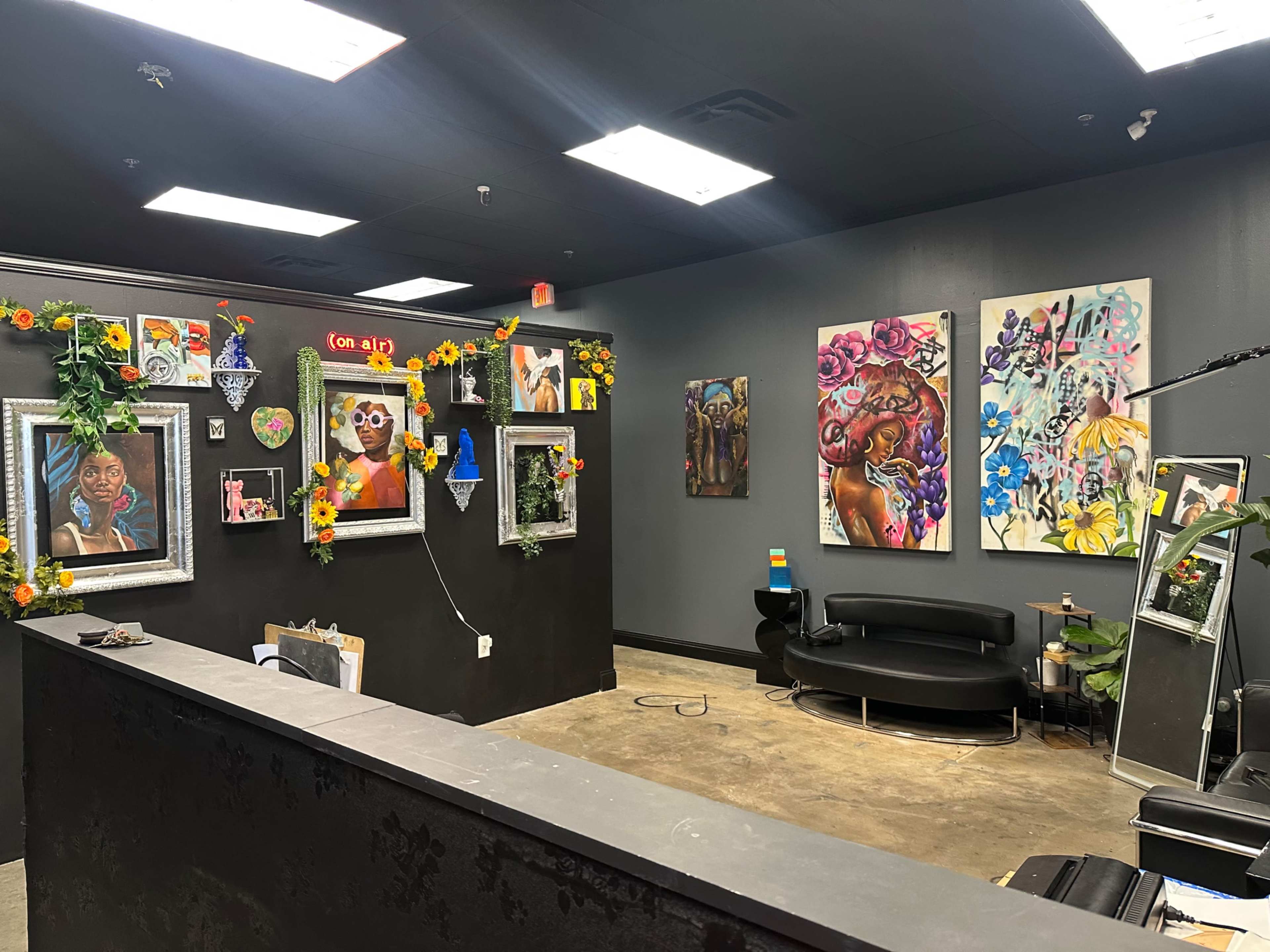 The image shows a vibrant art studio or gallery featuring colorful paintings on the walls, framed artwork, and decorative elements, including plants and flowers.