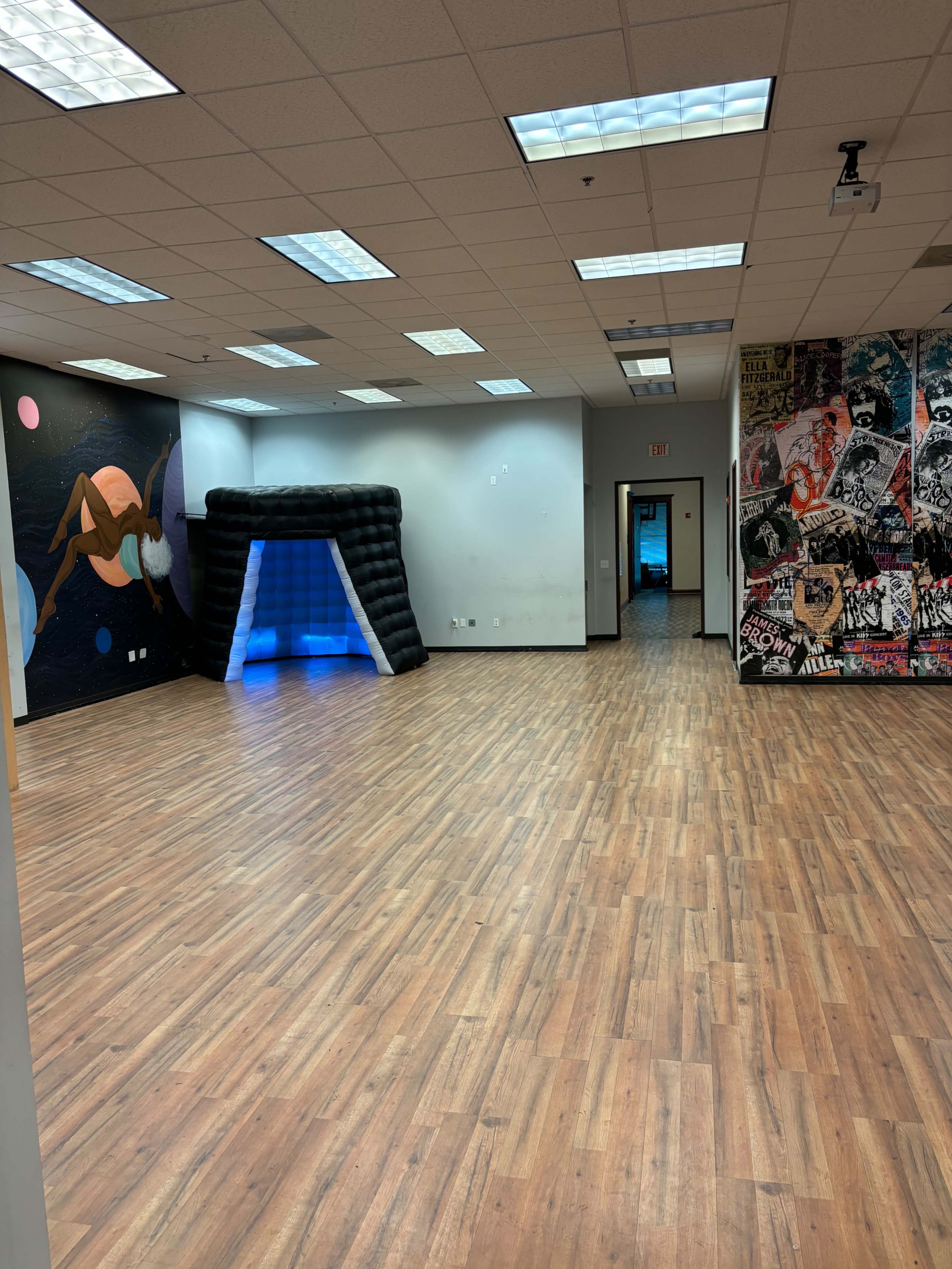 The image shows a spacious, empty room with wood flooring, featuring a large inflatable archway illuminated in blue and colorful wall murals.