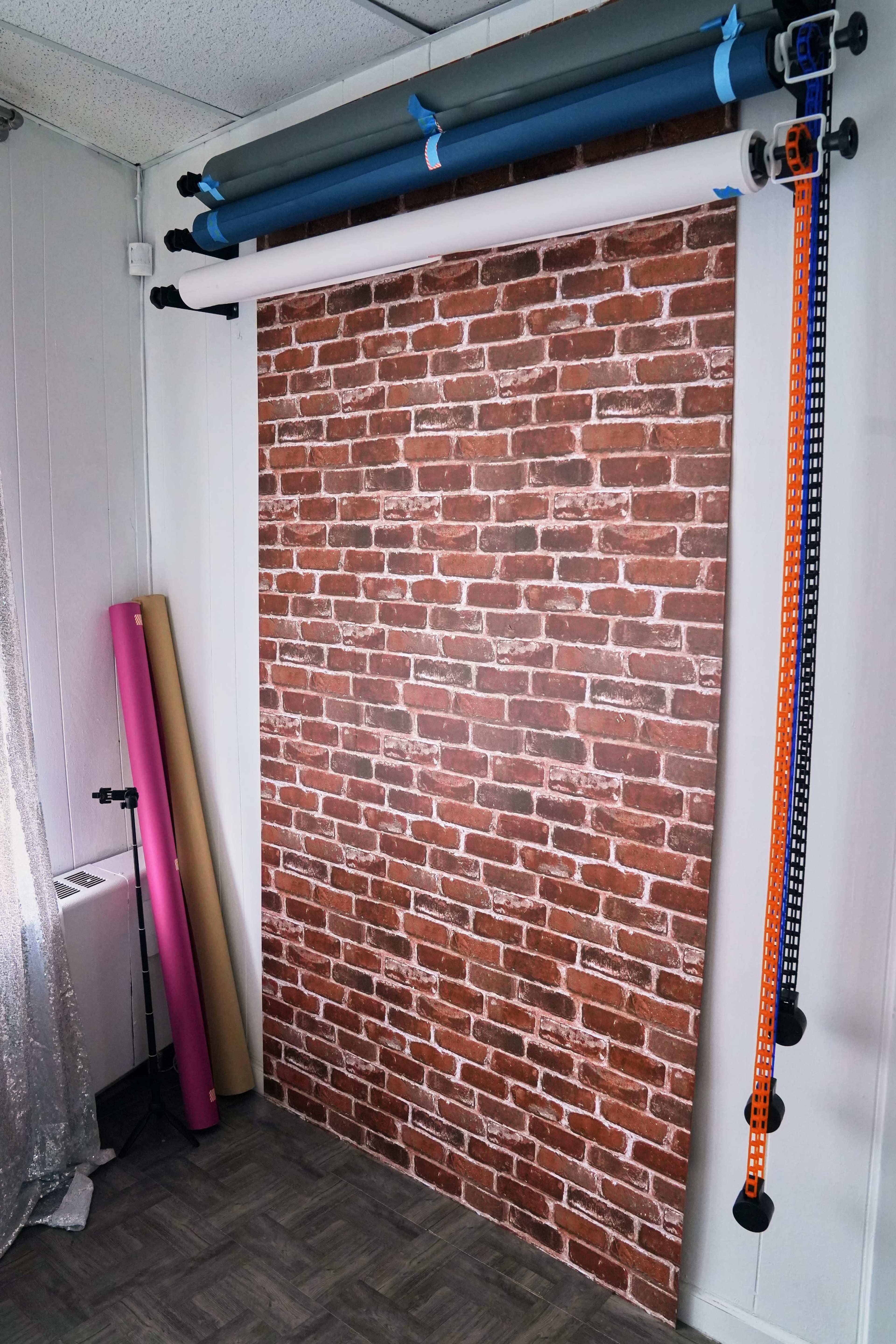 The image shows a photography setup featuring a brick-pattern backdrop, several rolls of backdrop paper, and a stand with colored straps in a room with gray flooring.