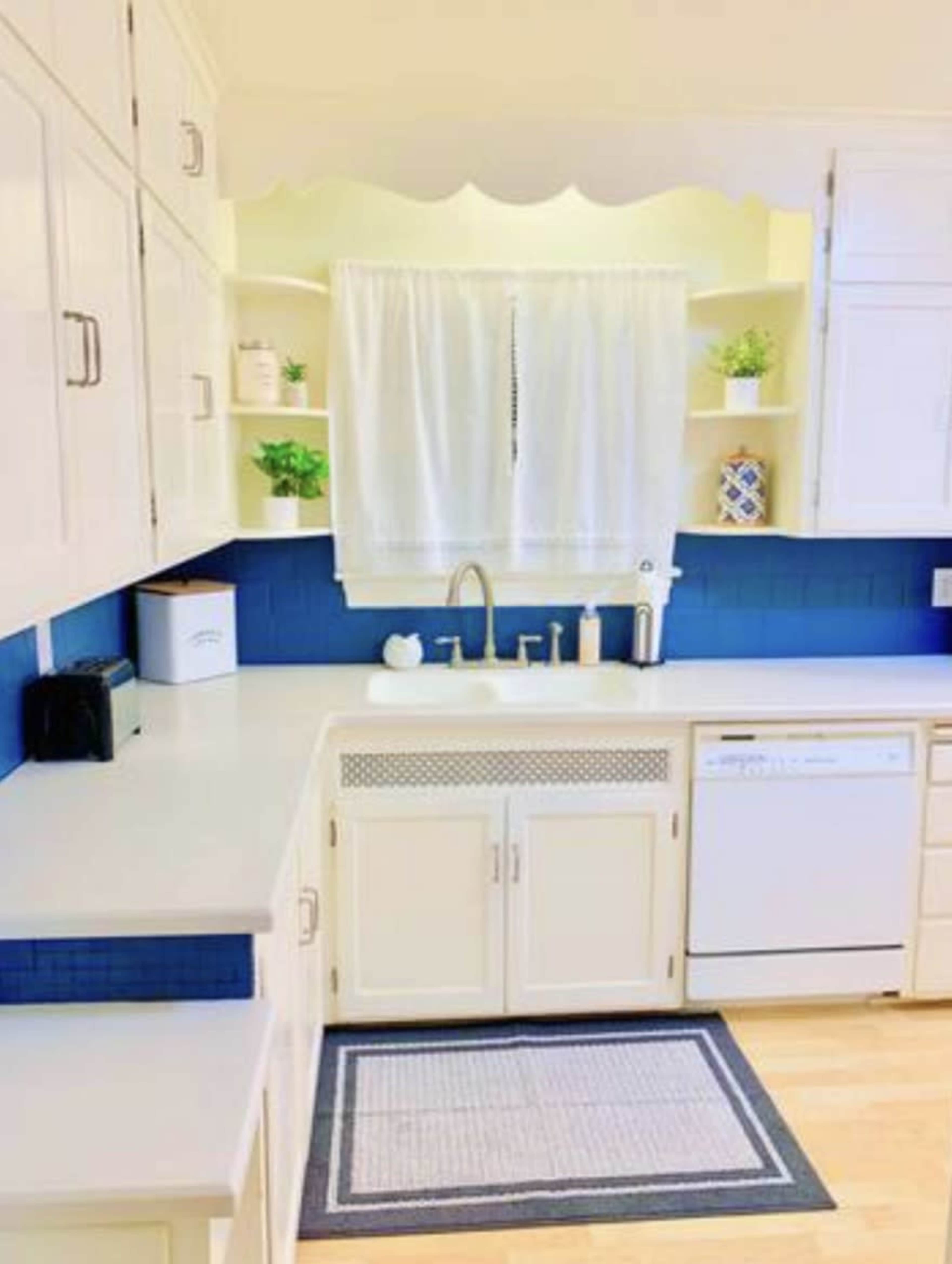 A bright kitchen features white cabinetry, a blue backsplash, and a window with a sheer curtain above the sink.