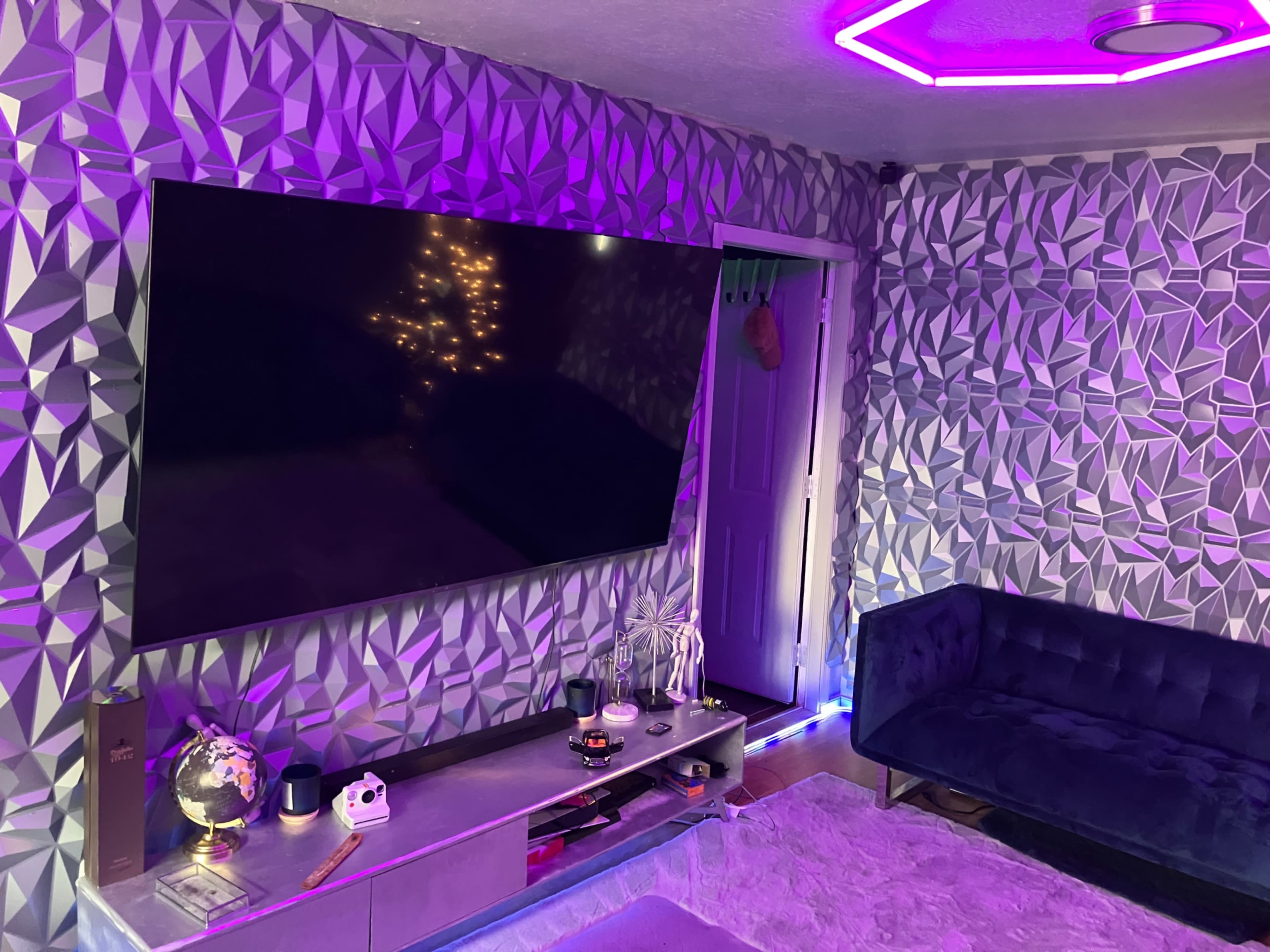 A modern living room features a large flat-screen TV mounted on a geometric-patterned wall, a dark blue sofa, and ambient purple lighting.