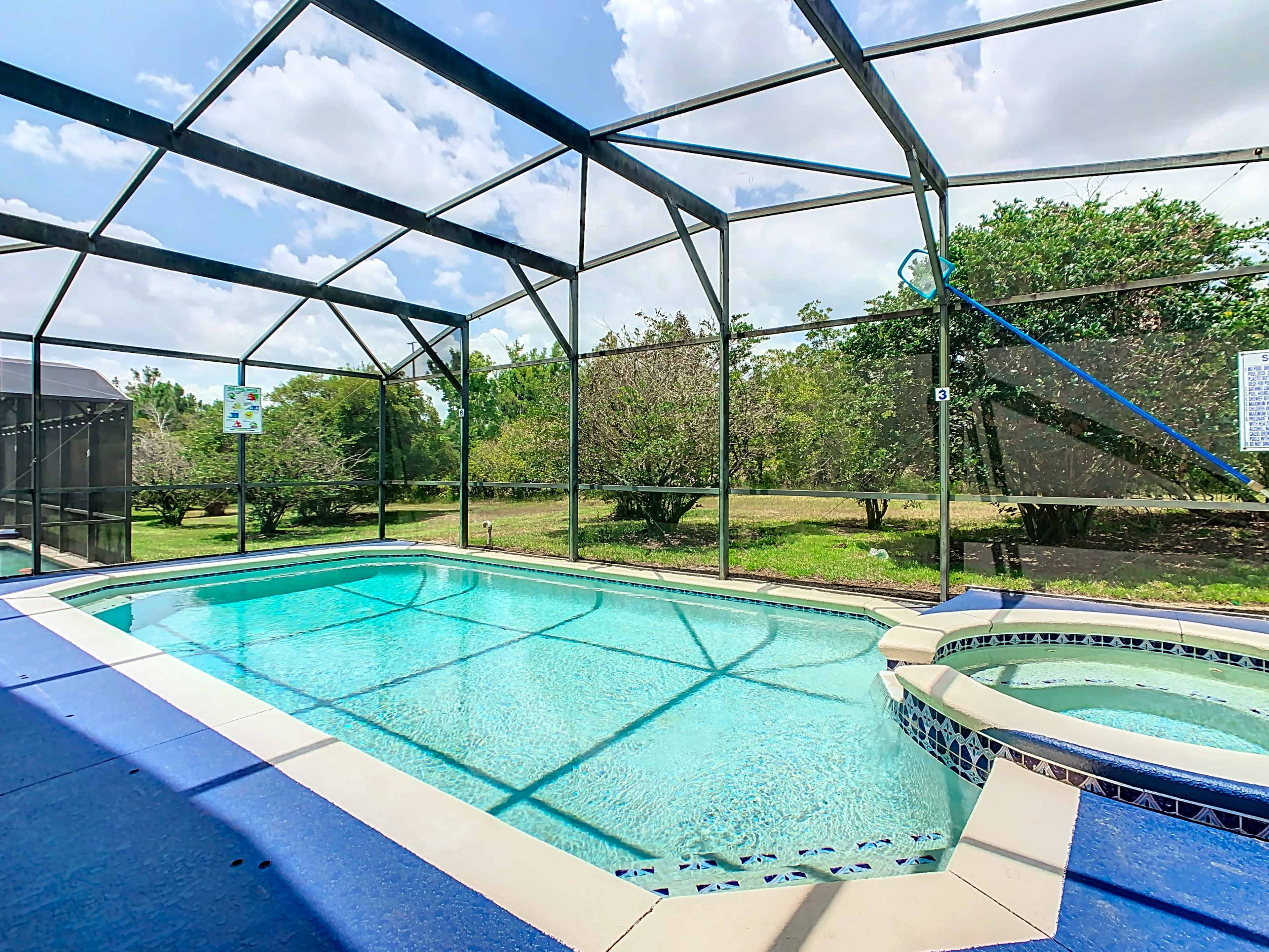 The image shows an enclosed swimming pool area with a clear blue pool and a hot tub, surrounded by greenery and a blue sky.