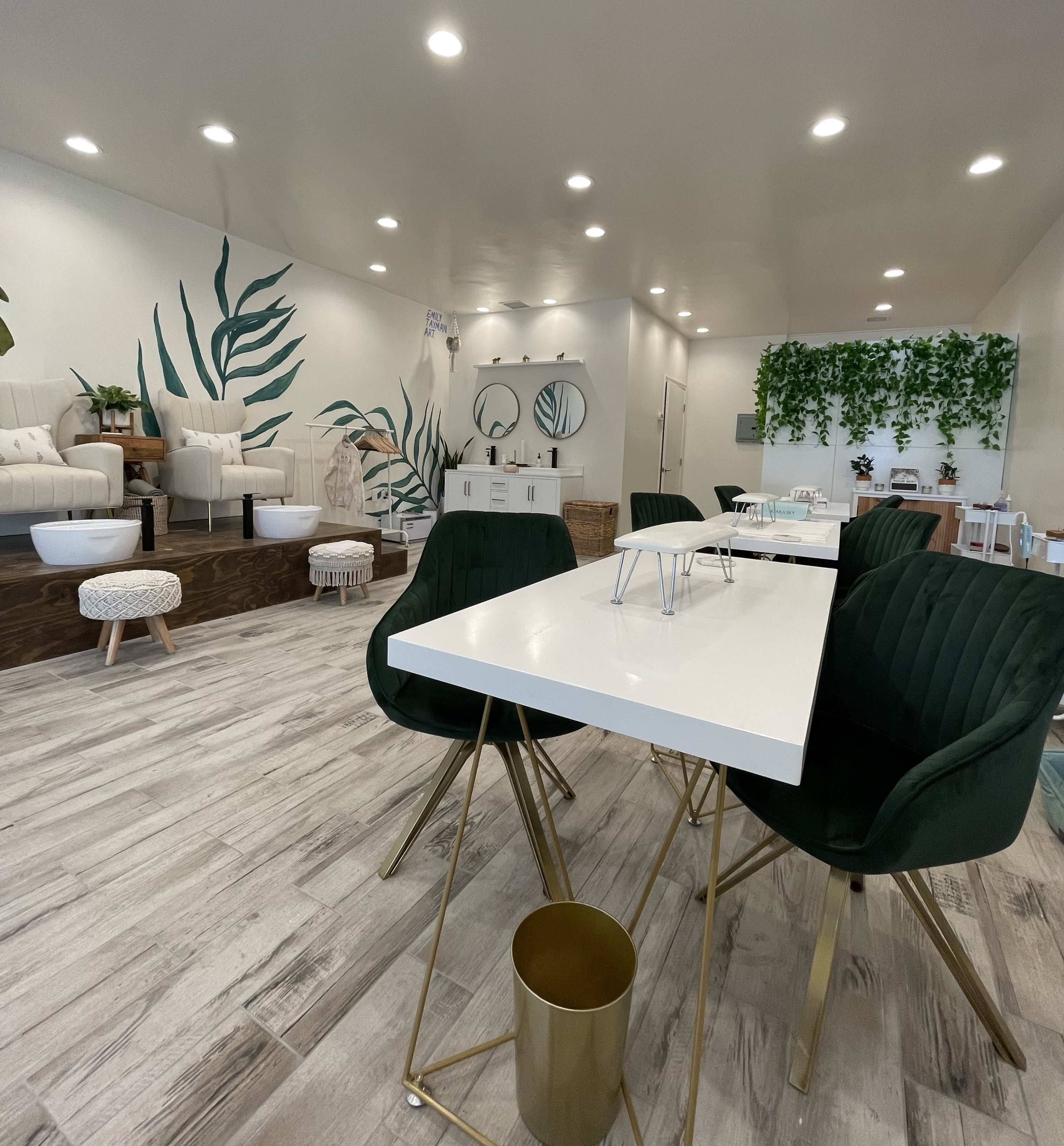 A modern, minimalist interior of a spa or salon featuring white tables, green chairs, and botanical wall decor.