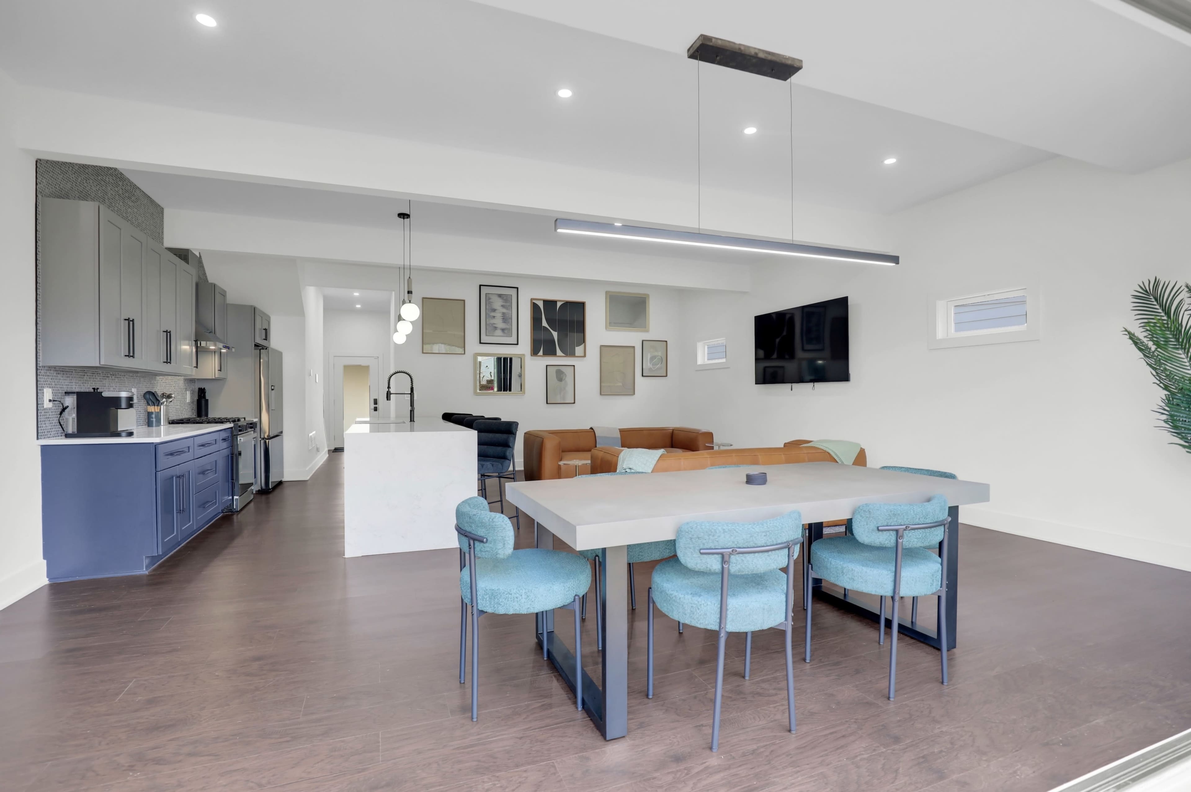 The image shows a modern open-concept living space with a dining area featuring a large table and blue chairs, adjacent to a kitchen with gray cabinets and a living room with a television.