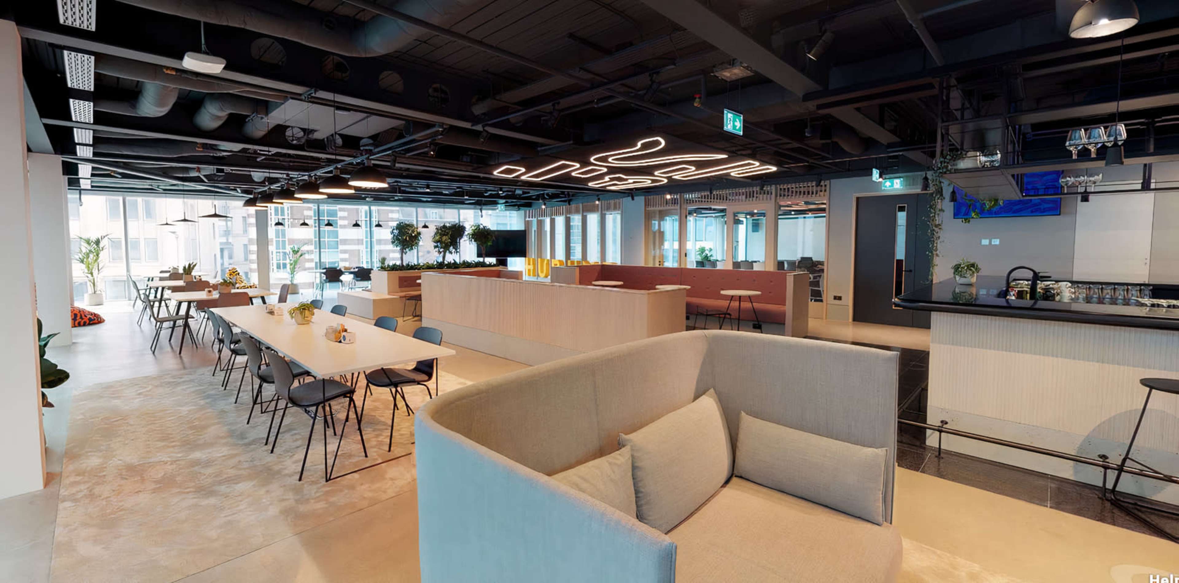 The image shows a modern office space with an open layout featuring seating areas, dining tables, and a large counter.