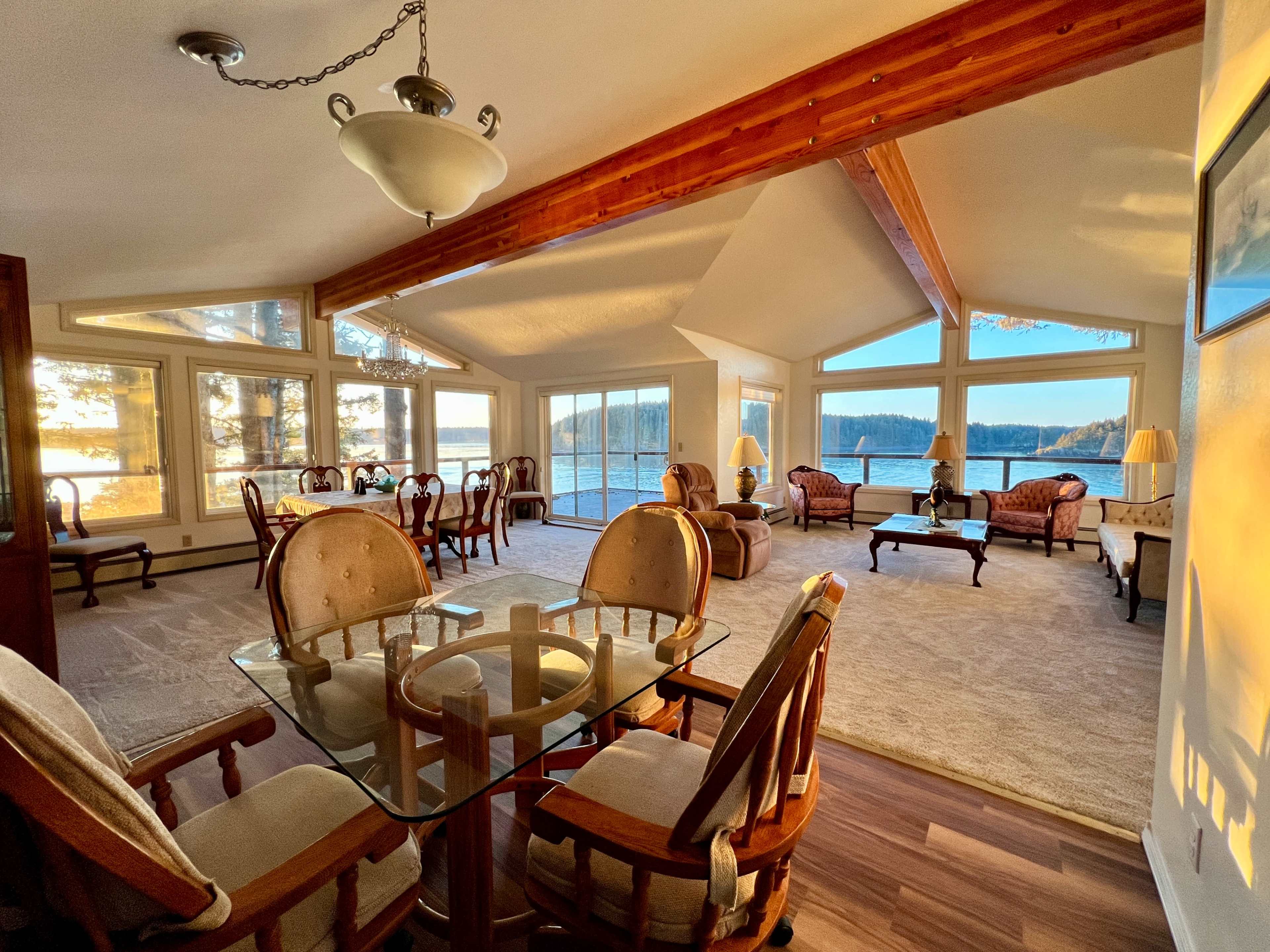 The image shows a spacious living area with large windows allowing natural light, furnished with comfortable seating and a dining table overlooking a lake.