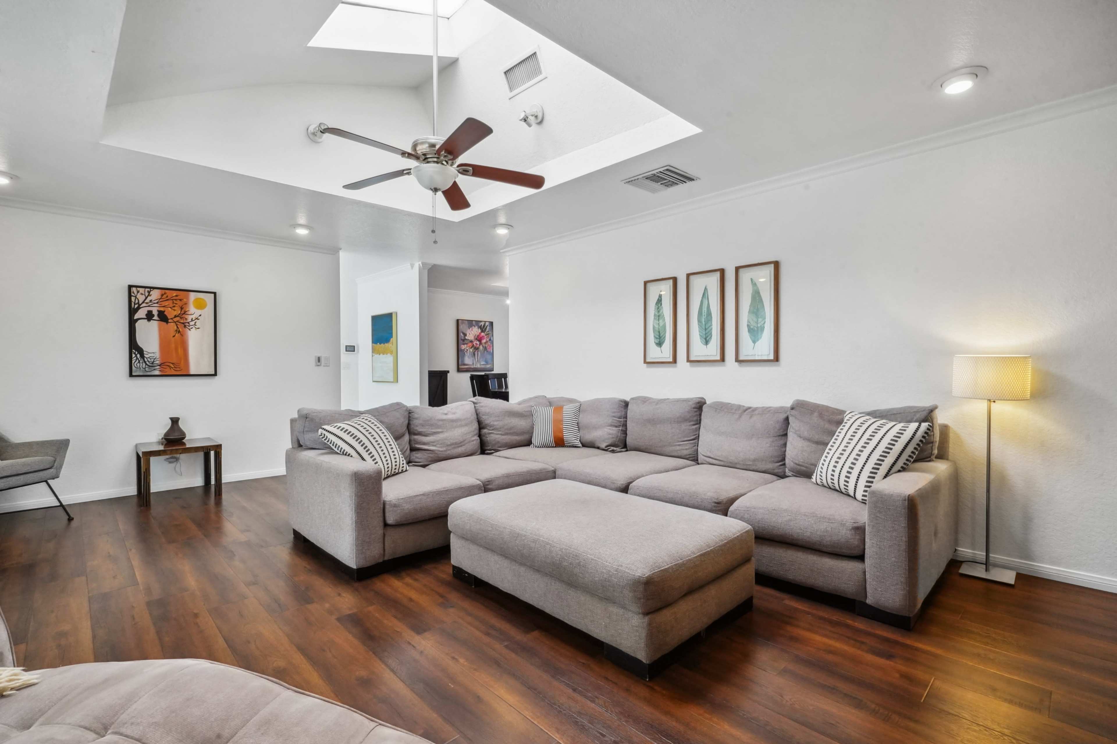 The living room features a large sectional sofa, a coffee table, and artwork on the walls, with natural light streaming through a skylight.