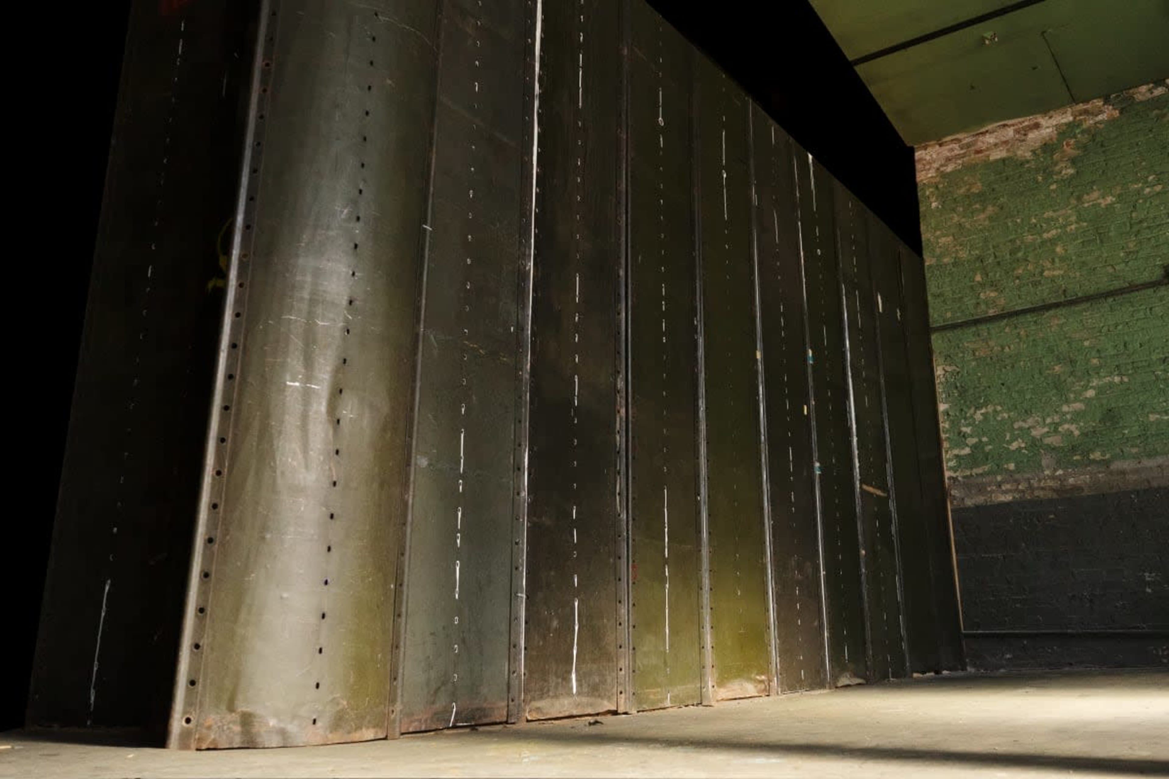 The image shows a series of large, dark metal panels arranged vertically against a textured green wall in an industrial setting.