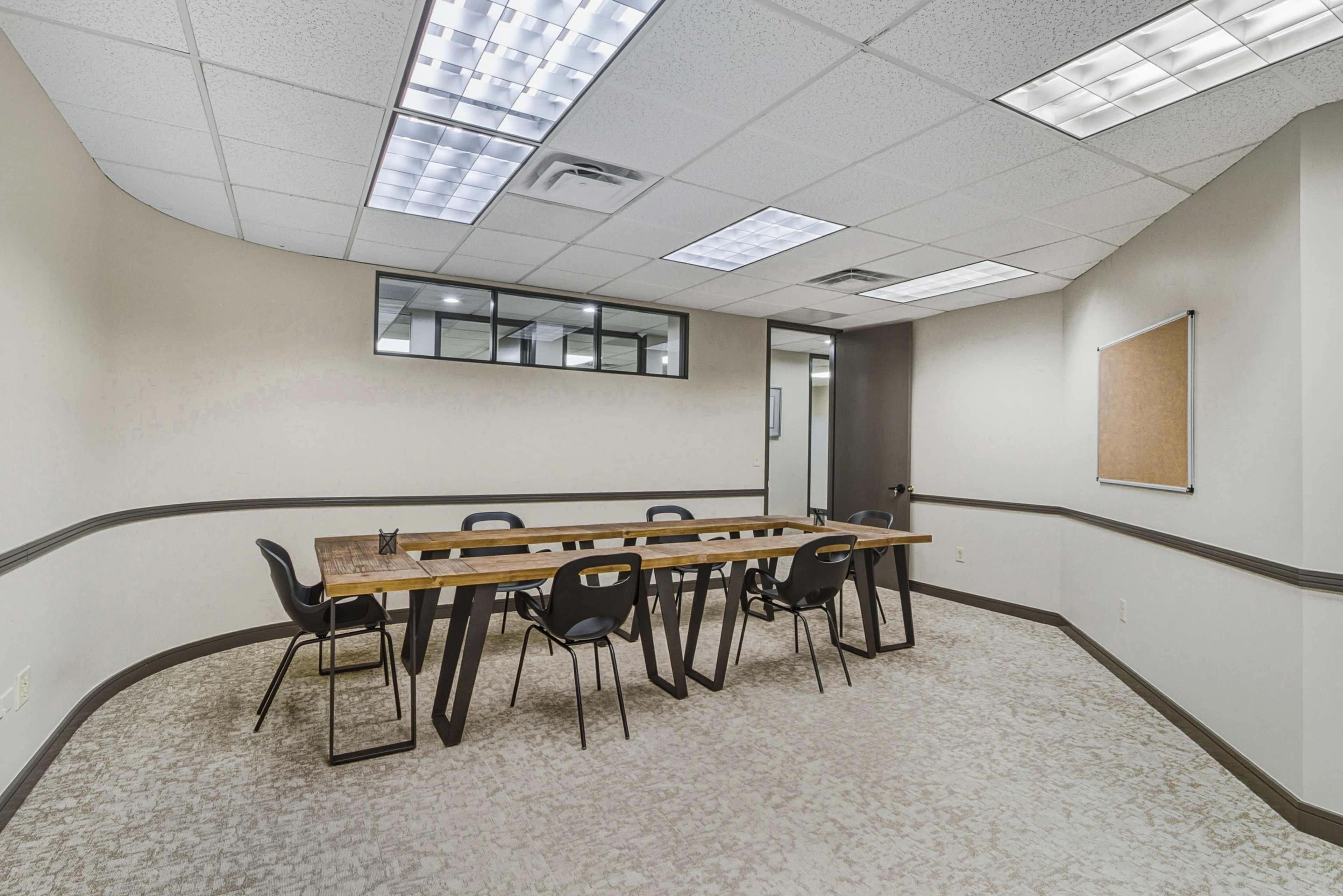 The image shows a modern conference room featuring a long wooden table surrounded by black chairs, with large windows and a bulletin board on one wall.