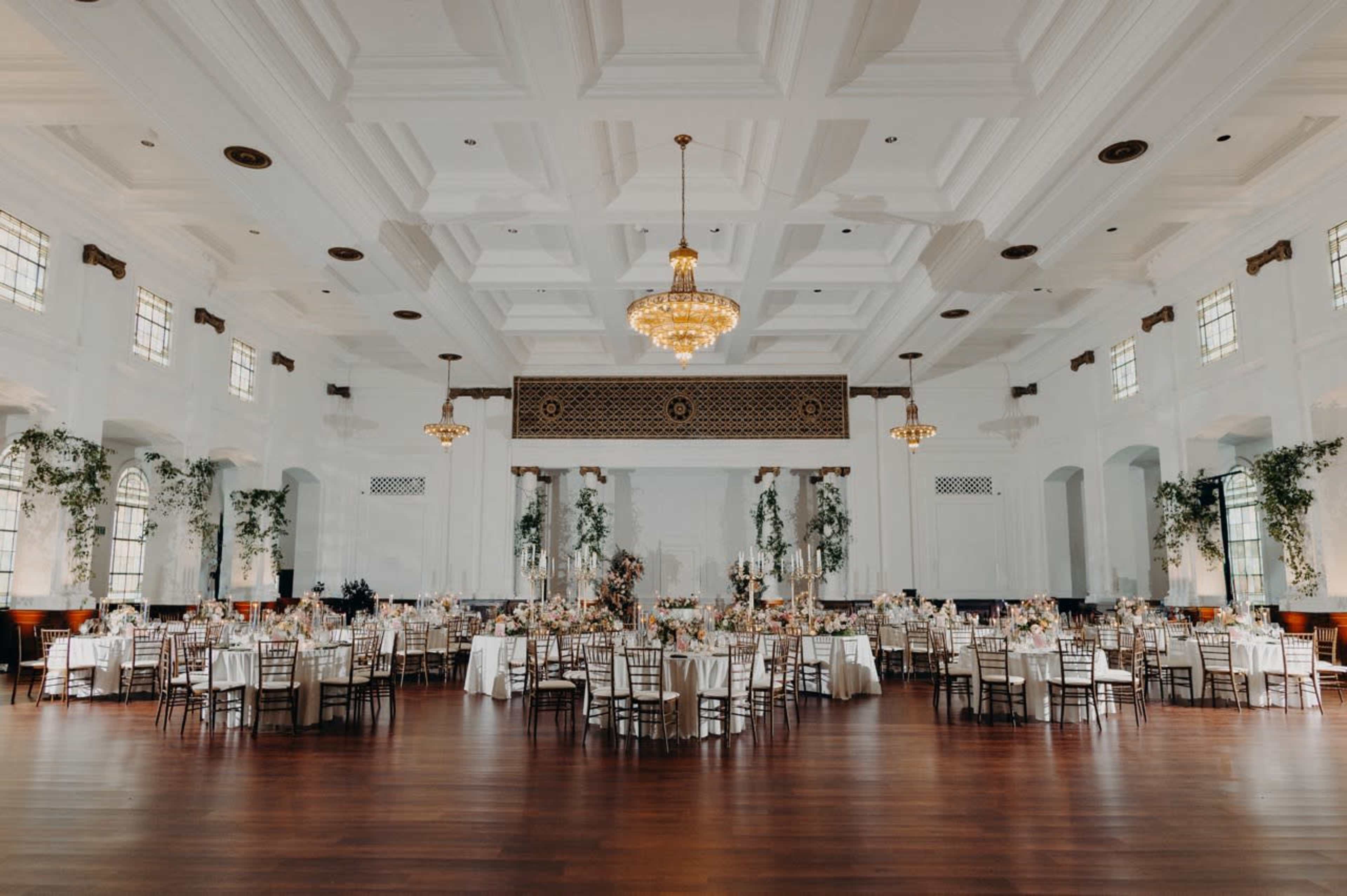 The image shows a grand banquet hall set up with round tables adorned with white tablecloths and floral centerpieces, surrounded by elegant decorations and a large chandelier overhead.