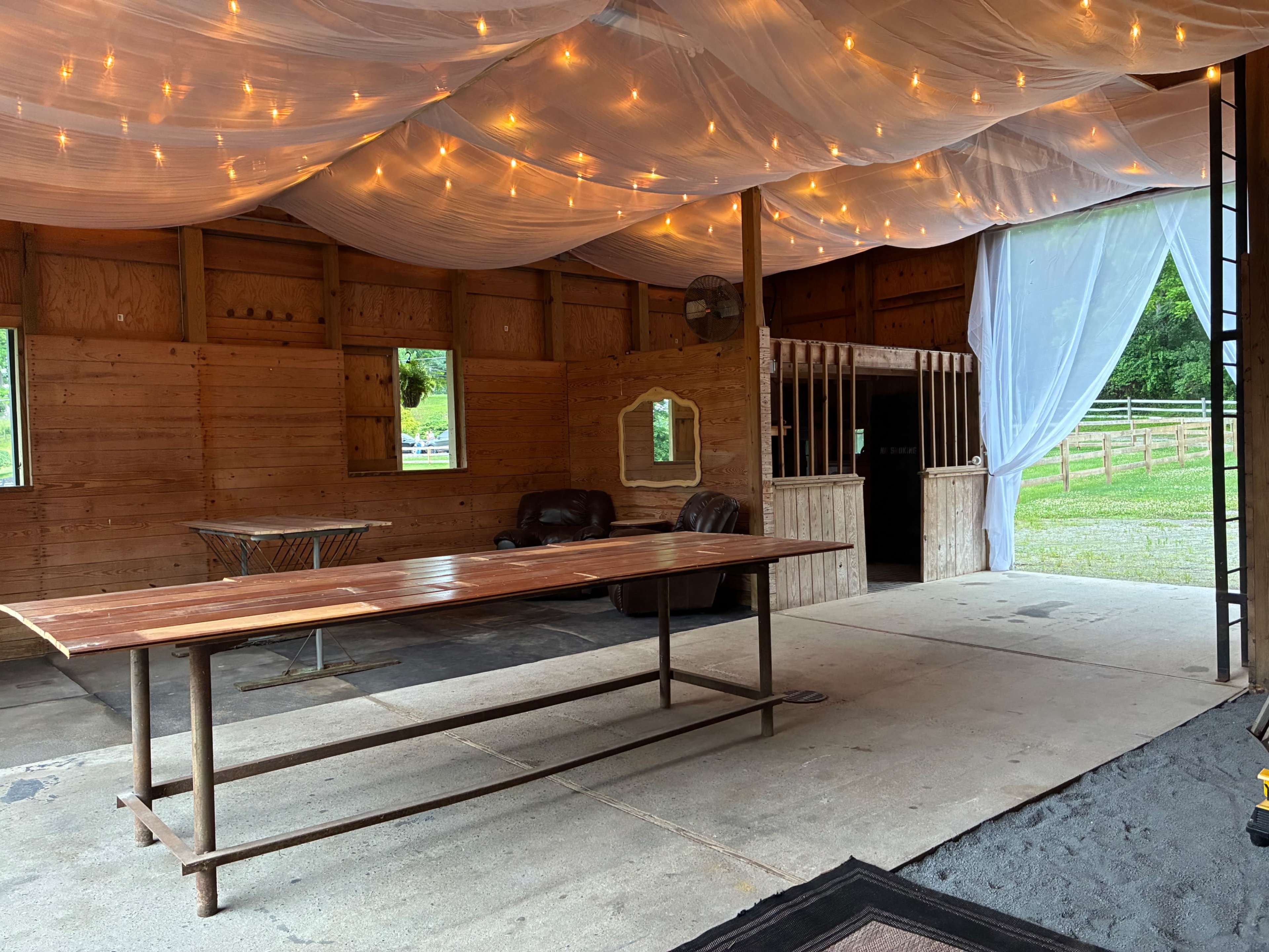 The image shows an indoor space with a large wooden table, two leather chairs, and draped fabric overhead illuminated by string lights, with a wooden partition visible in the background.