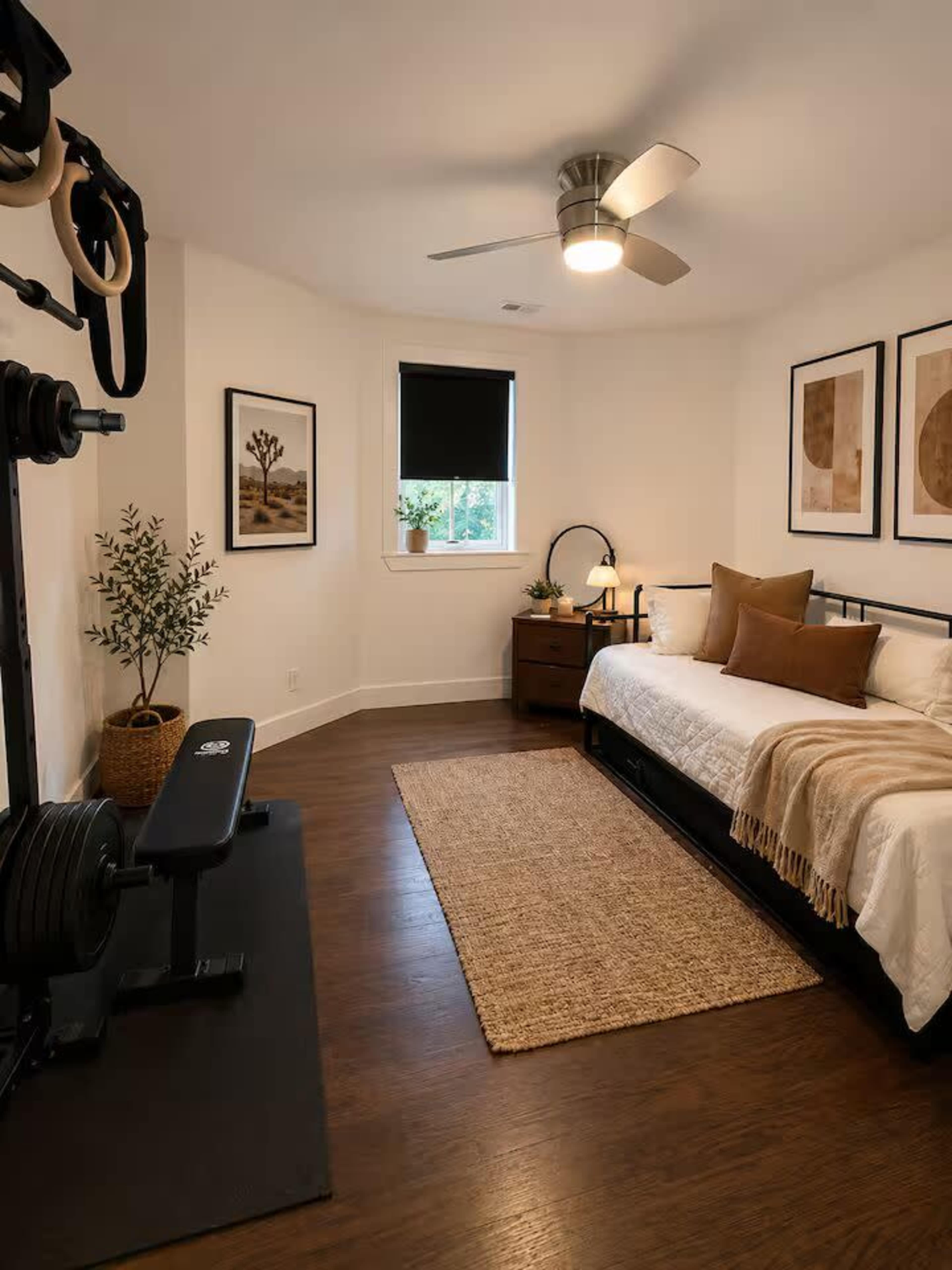 The image shows a small bedroom featuring a daybed with decorative pillows, a wooden nightstand, a workout bench with weights, and framed art on the walls.