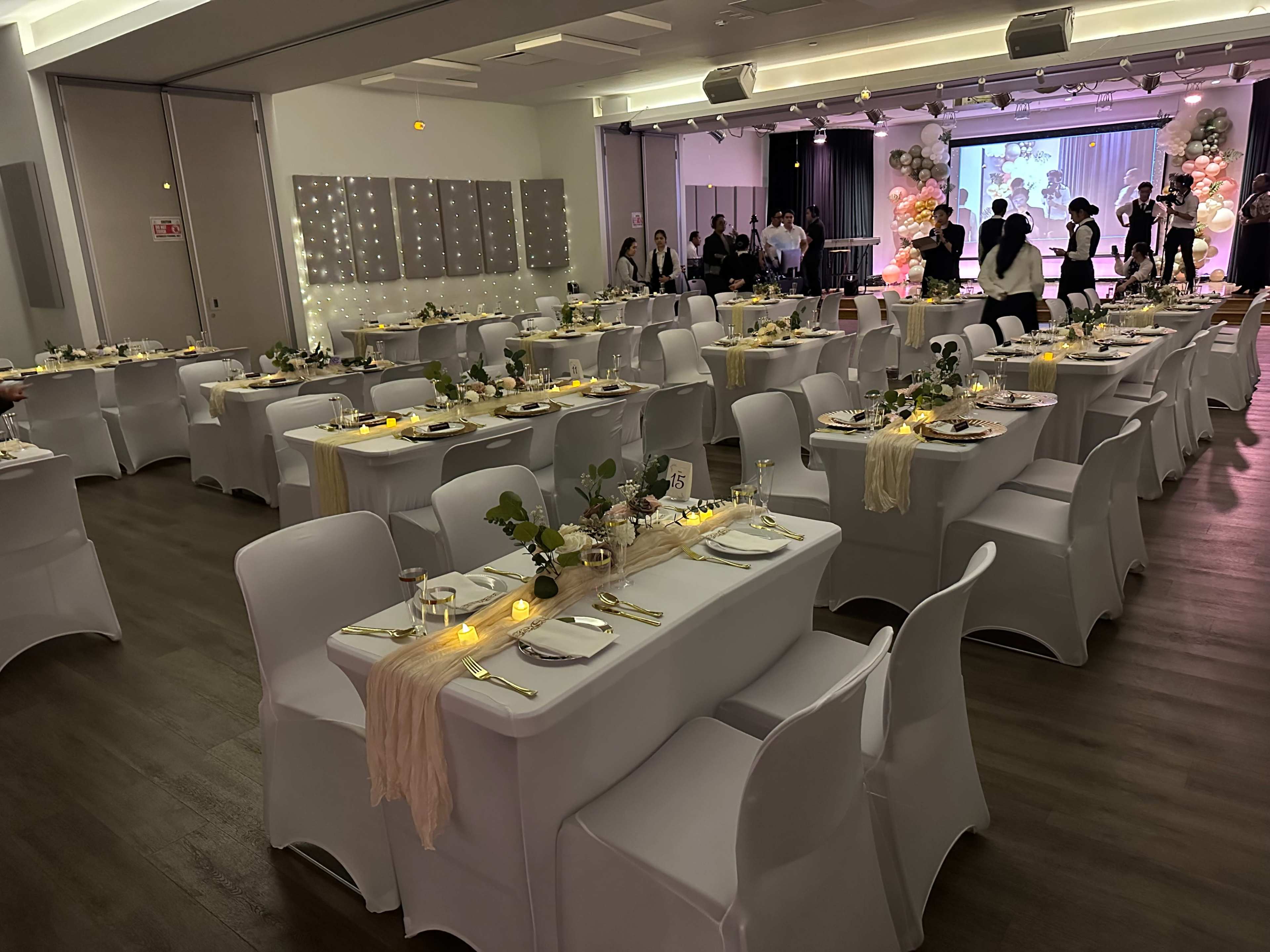 The image depicts a banquet hall set for an event, featuring rows of elegantly arranged tables covered with white tablecloths, centerpieces of greenery, and dim lighting against a backdrop of a stage with a projector screen.