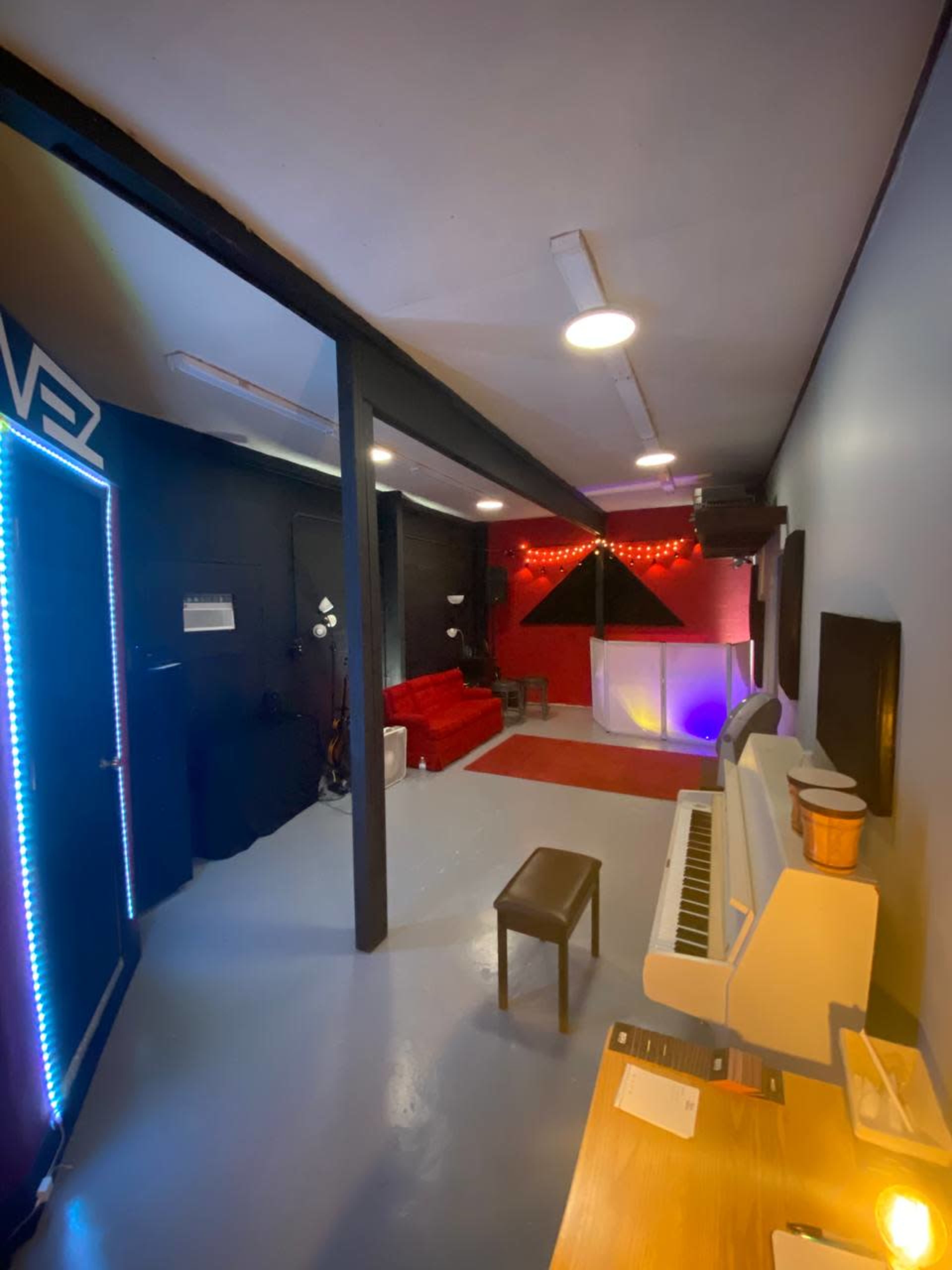 A small recording studio featuring a white piano, a black bench, red couches, and geometric art illuminated by colored lights.