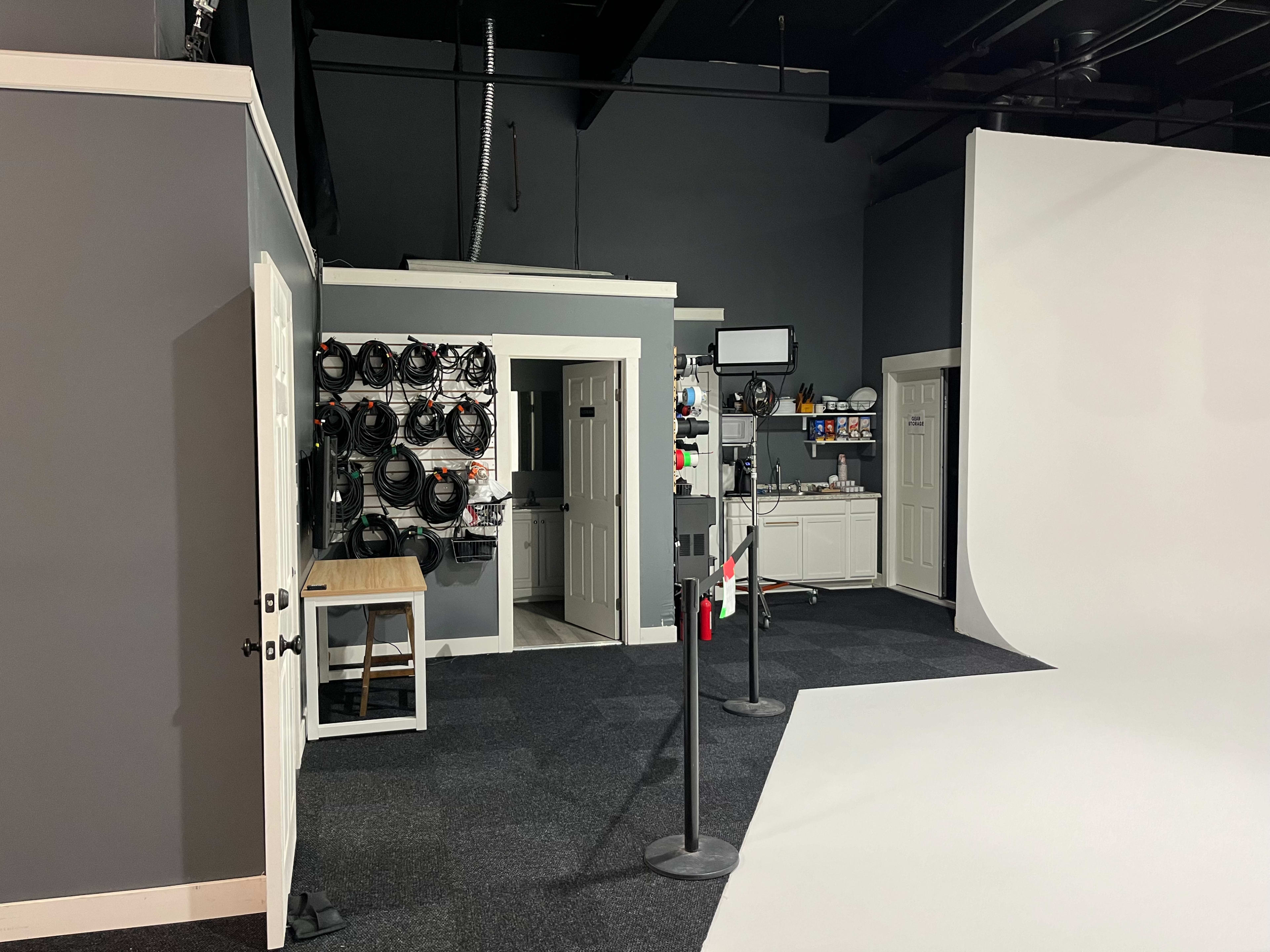 The image shows a photography studio featuring a gray wall, a blank white backdrop, and various equipment organized along one side.