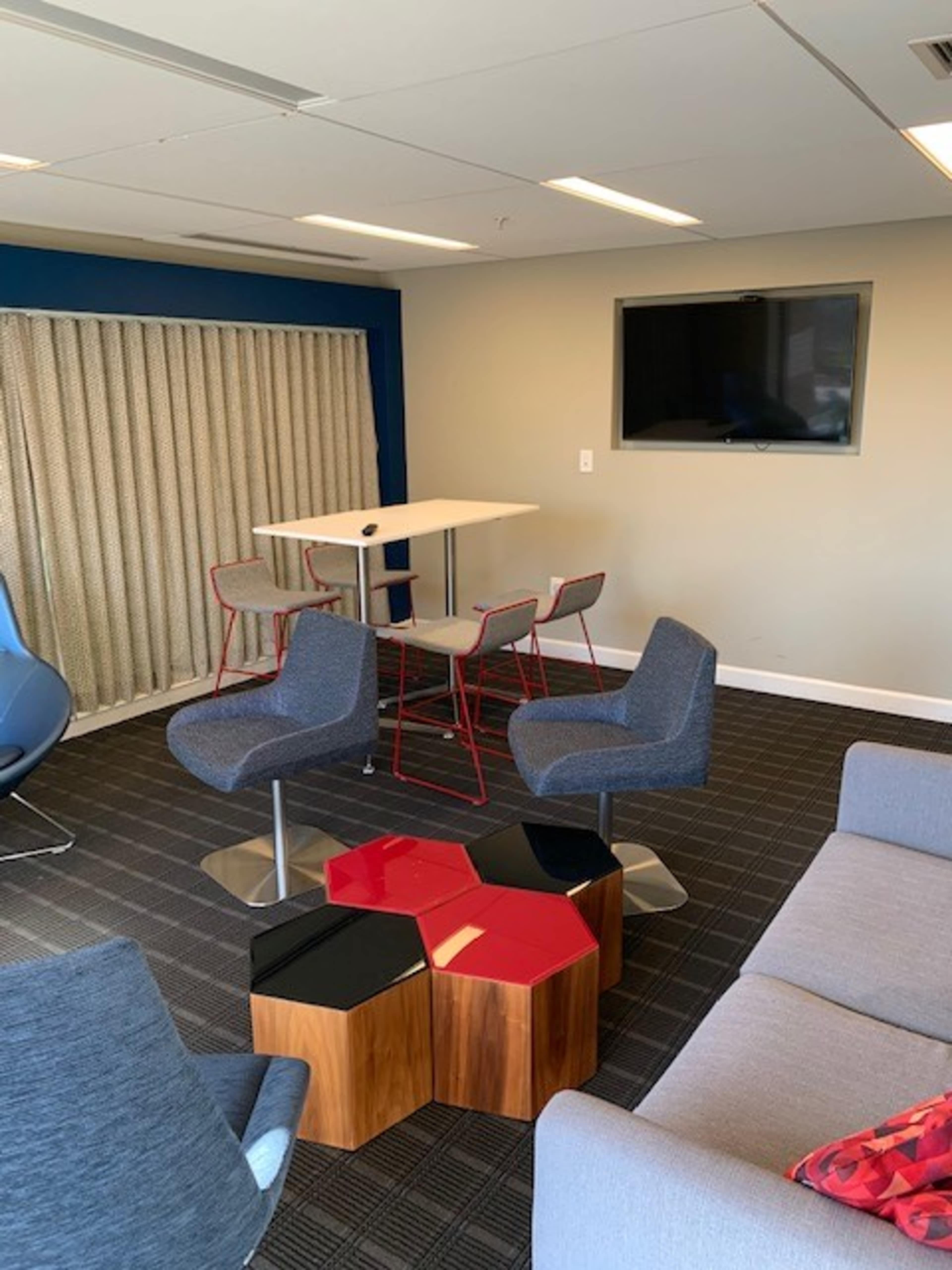 A modern lounge area features a gray sofa, hexagonal wooden coffee table, blue chairs, and a high table with barstools, along with a wall-mounted television.
