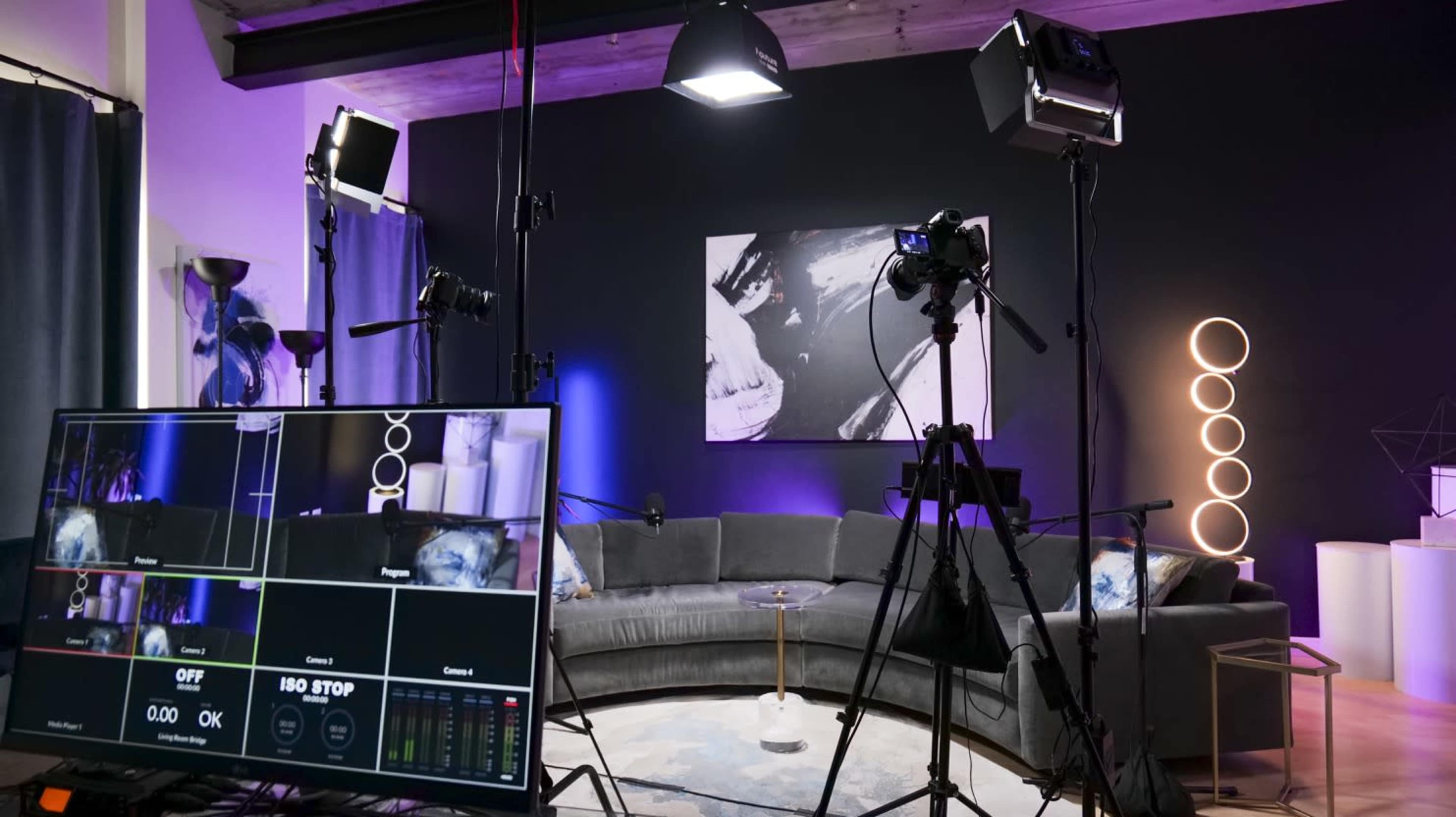 The image shows a studio setup for video production featuring a circular gray couch, professional lighting equipment, and a control monitor displaying various camera feeds.