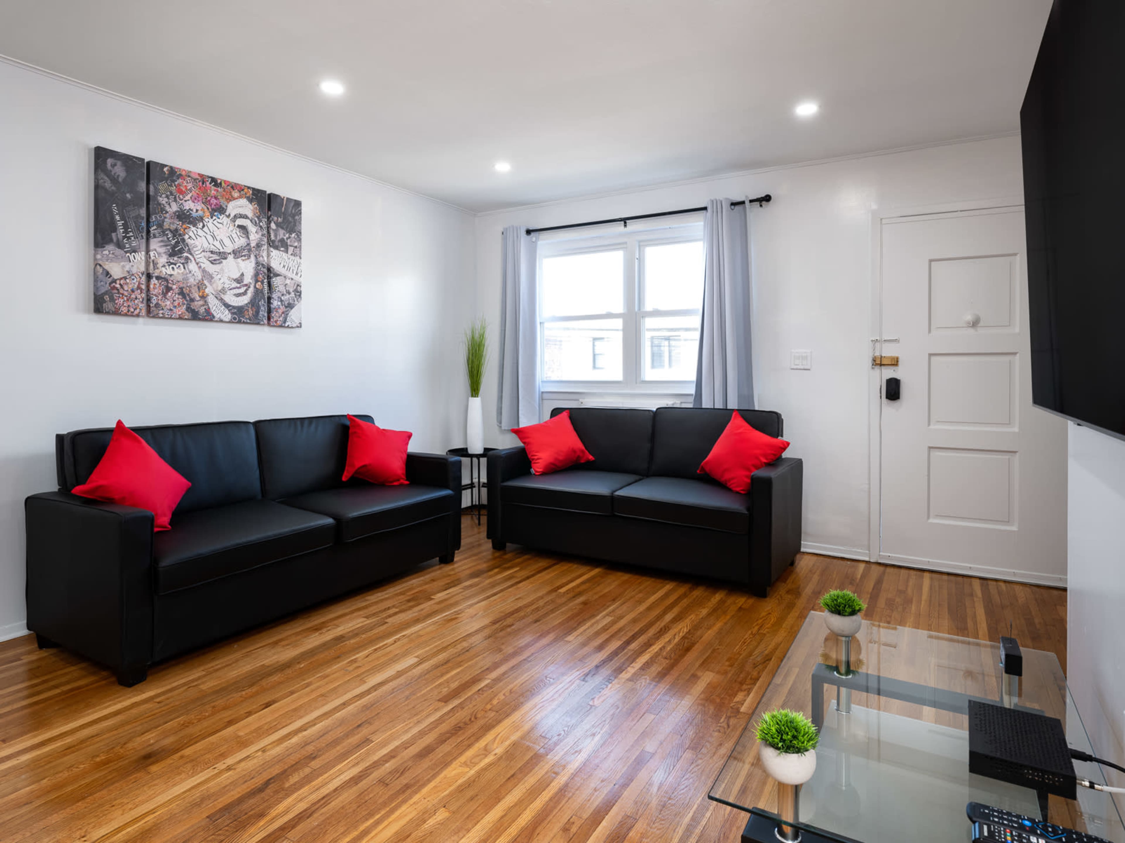 The living room features two black sofas with red pillows, a glass coffee table, and a wall-mounted television, illuminated by natural light from two windows.