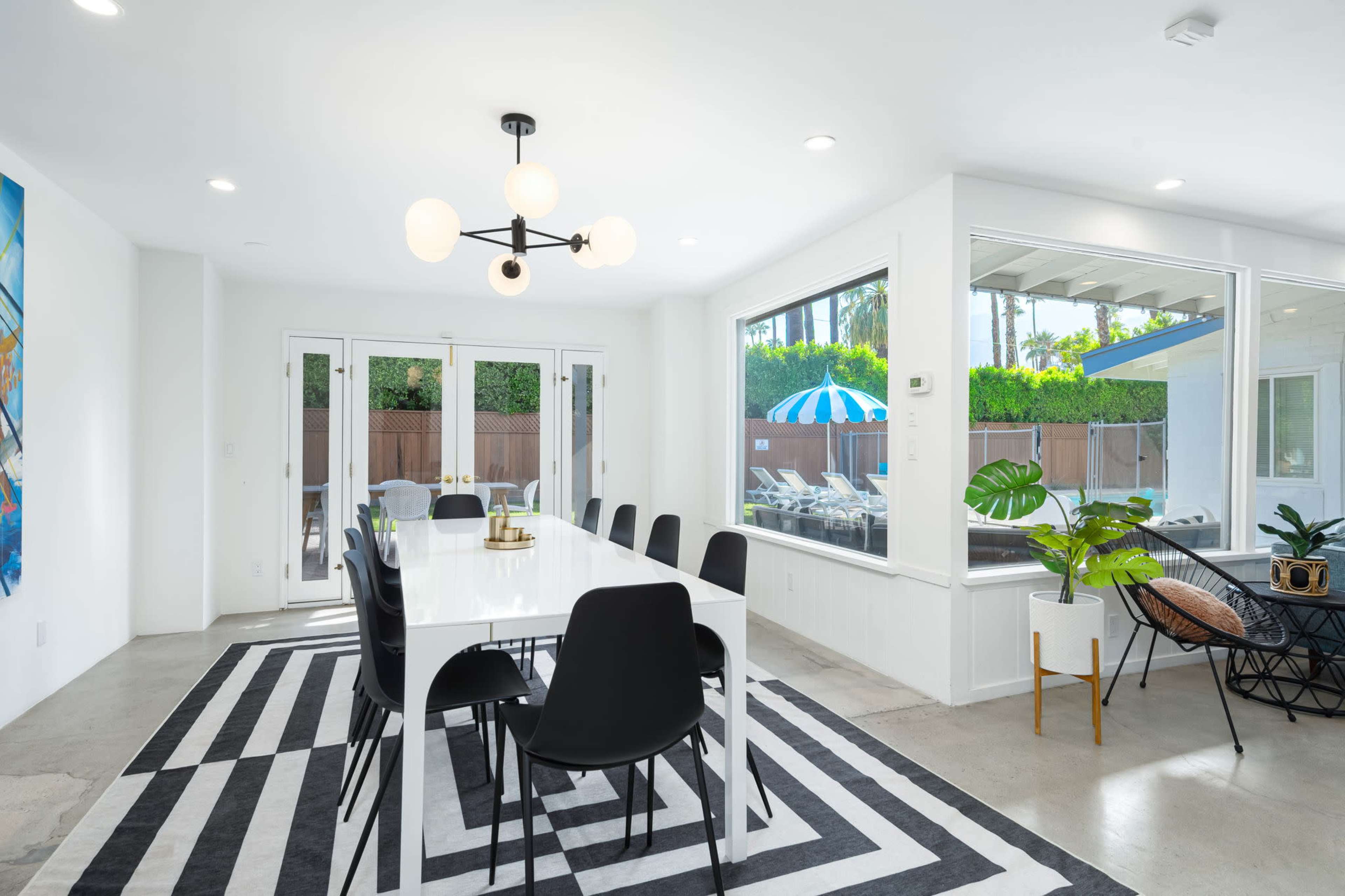 A dining area features a long white table surrounded by black chairs on a striped rug, with large windows providing a view of an outdoor space with loungers and a blue striped umbrella.