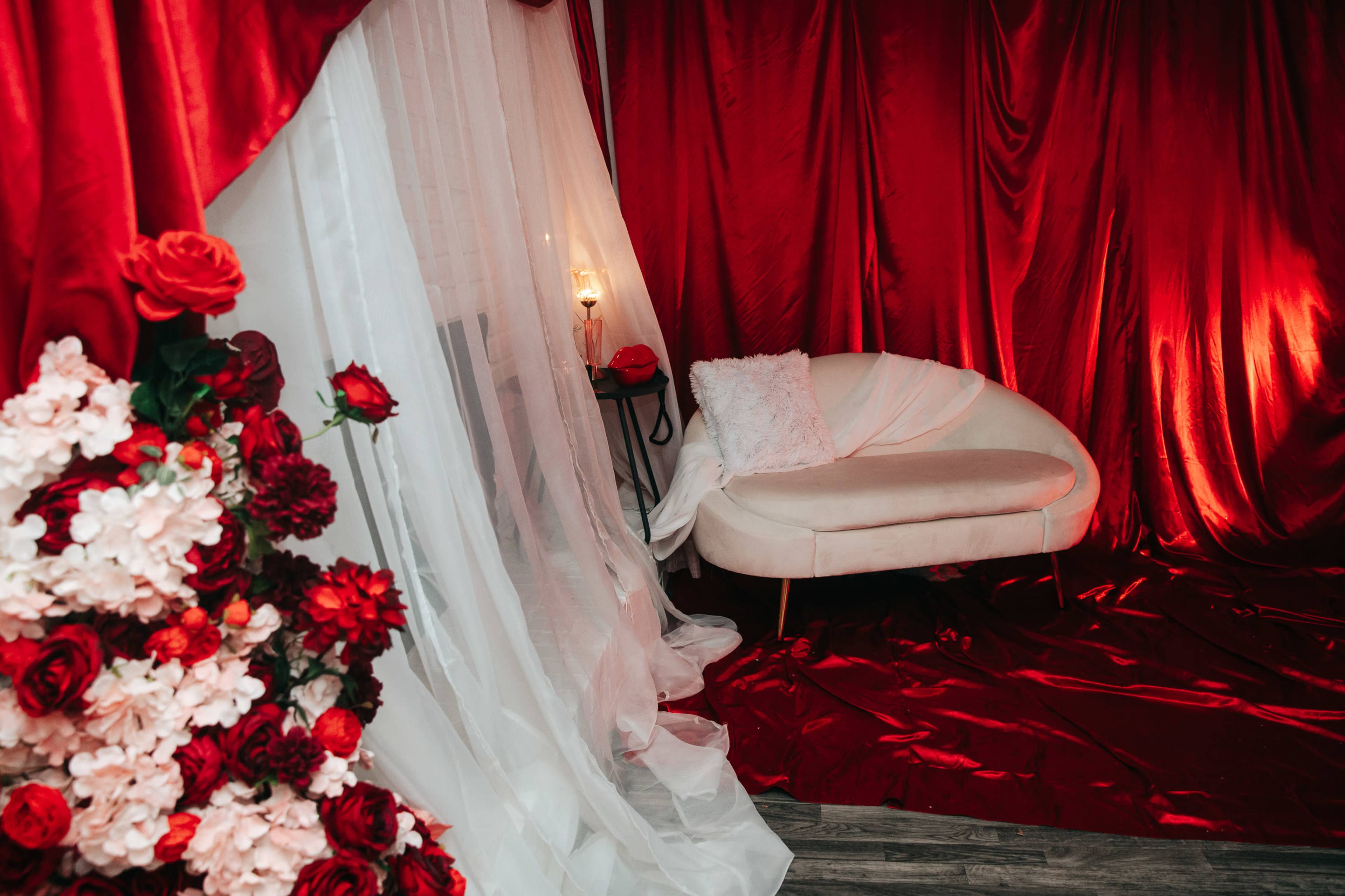 A red-themed room features a white curved couch surrounded by draped fabric and floral arrangements.