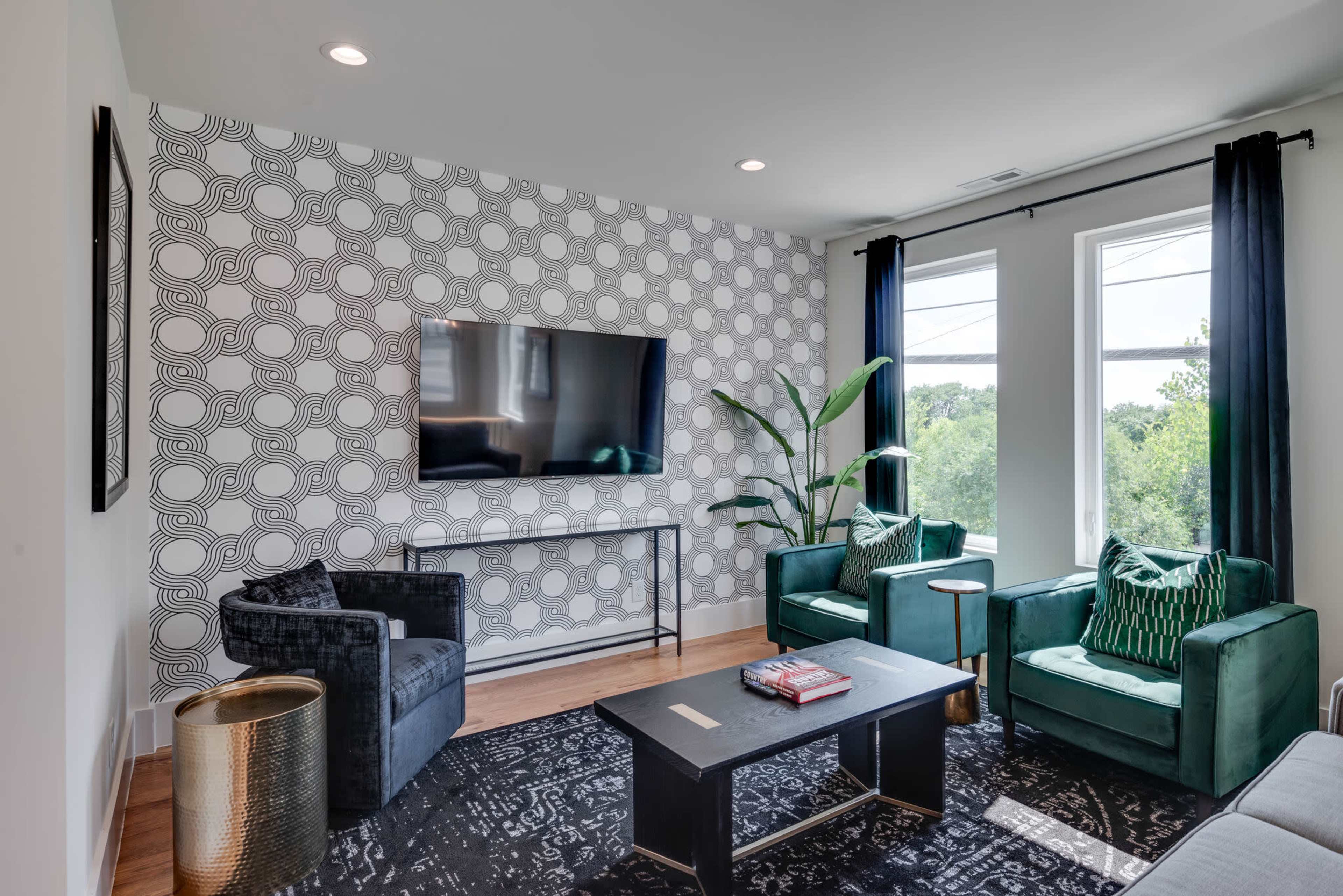 A modern living room features two green chairs, a dark coffee table, a large television on the wall, and large windows with a view of greenery outside.