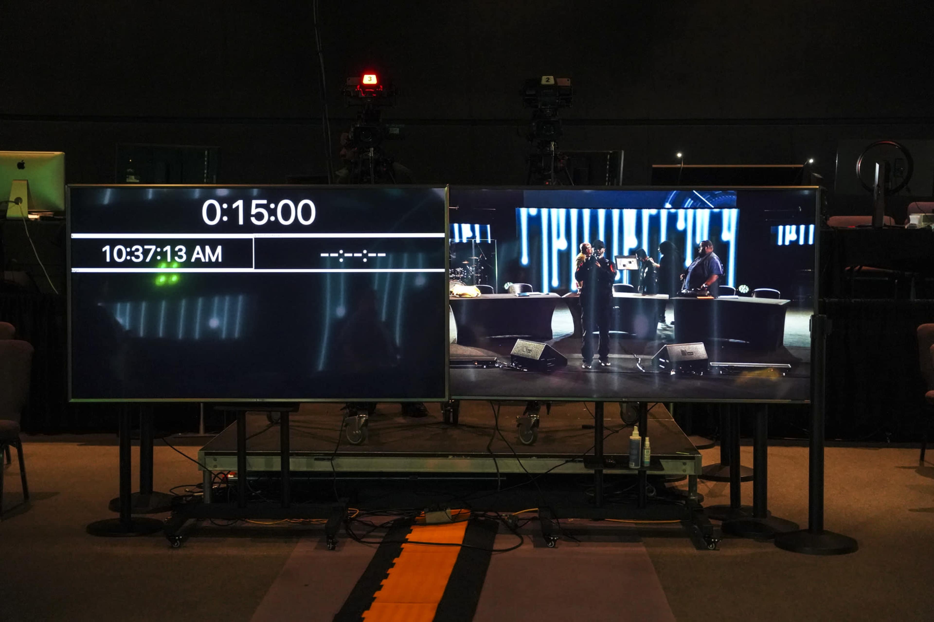 The image shows two large screens in a studio, one displaying a countdown timer and the other showing a live performance on stage.