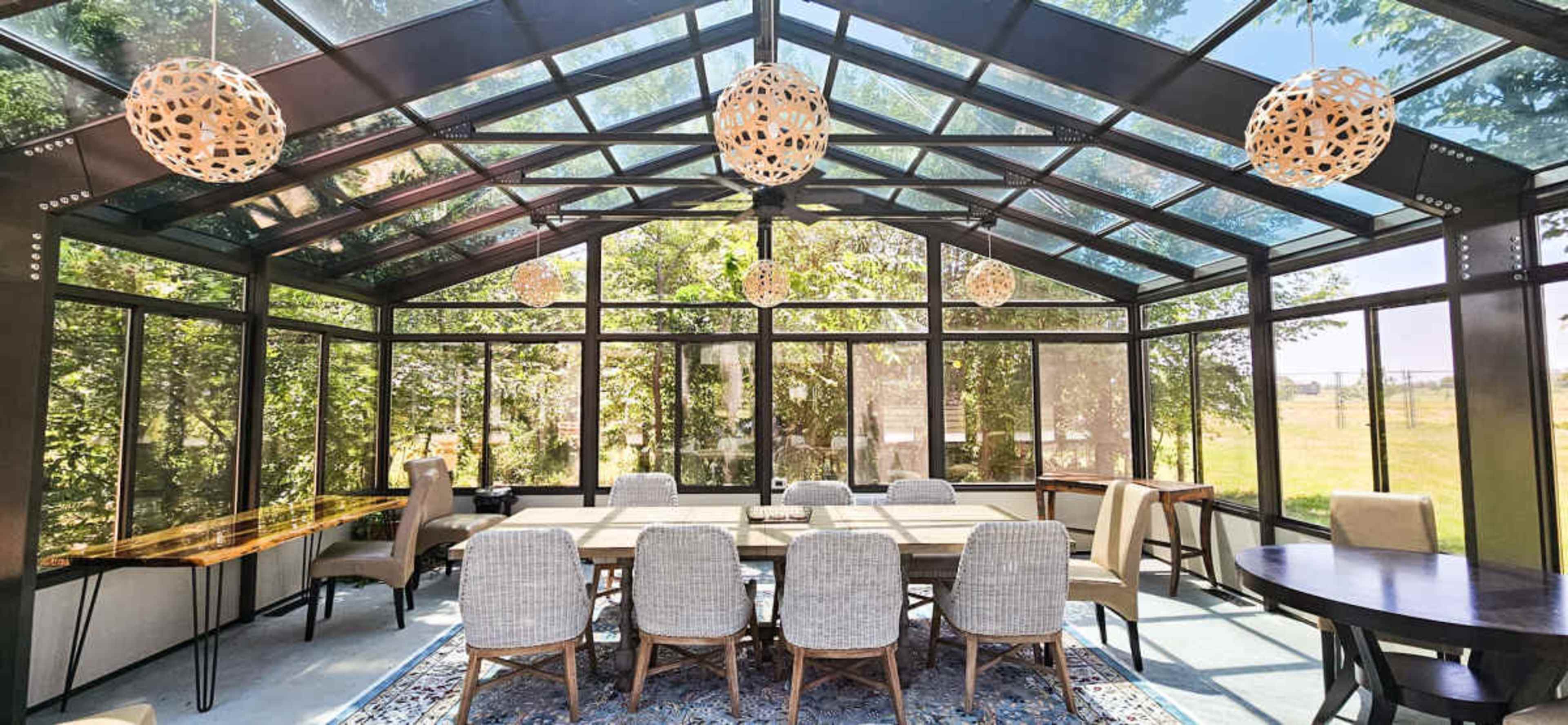 A spacious sunroom features a glass ceiling, multiple pendant lights, and a large dining table surrounded by upholstered chairs.