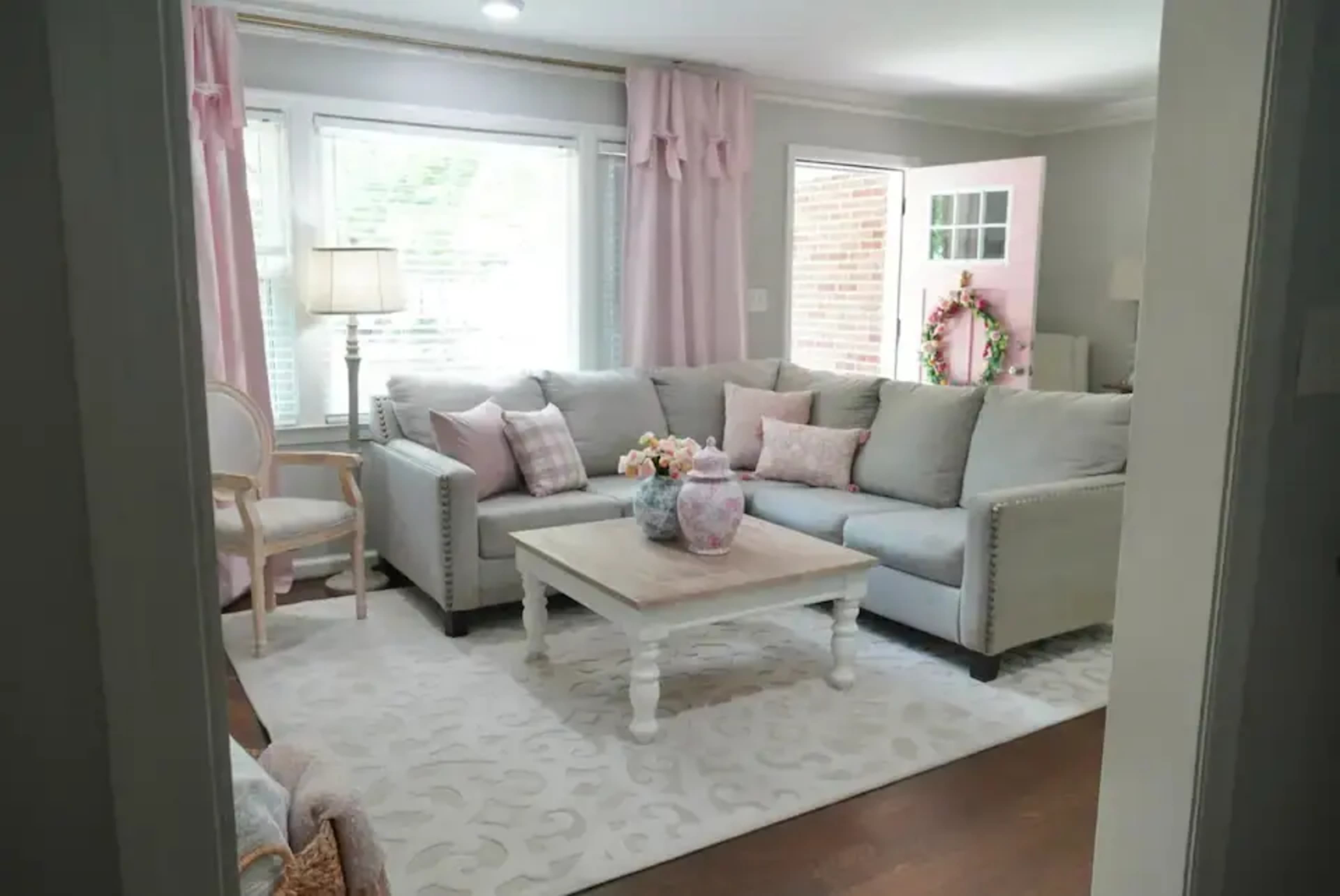 A living room features a gray sectional sofa, a white coffee table, and pastel-colored decor near large windows with pink curtains.