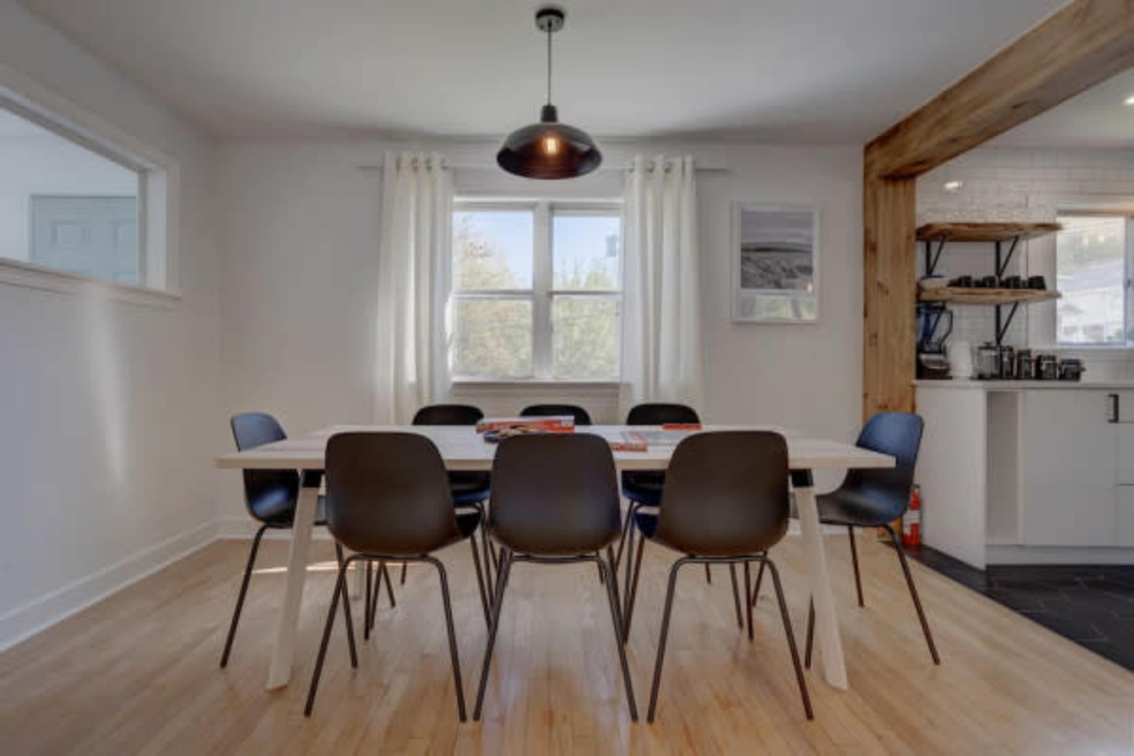 A modern dining room features a long white table surrounded by black chairs, with large windows letting in natural light.