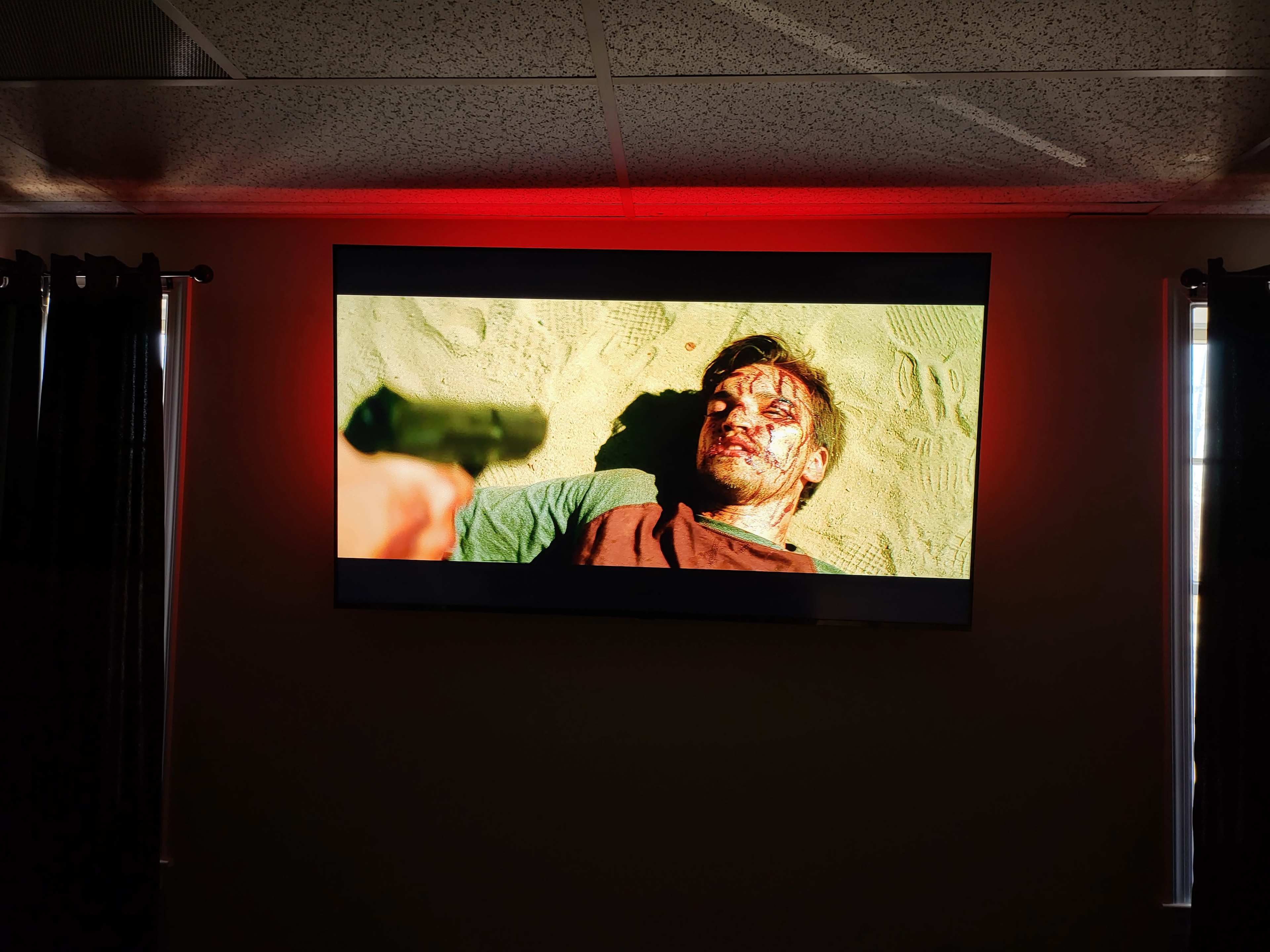 A man with blood on his face lies on the sand while another person aims a gun at him.