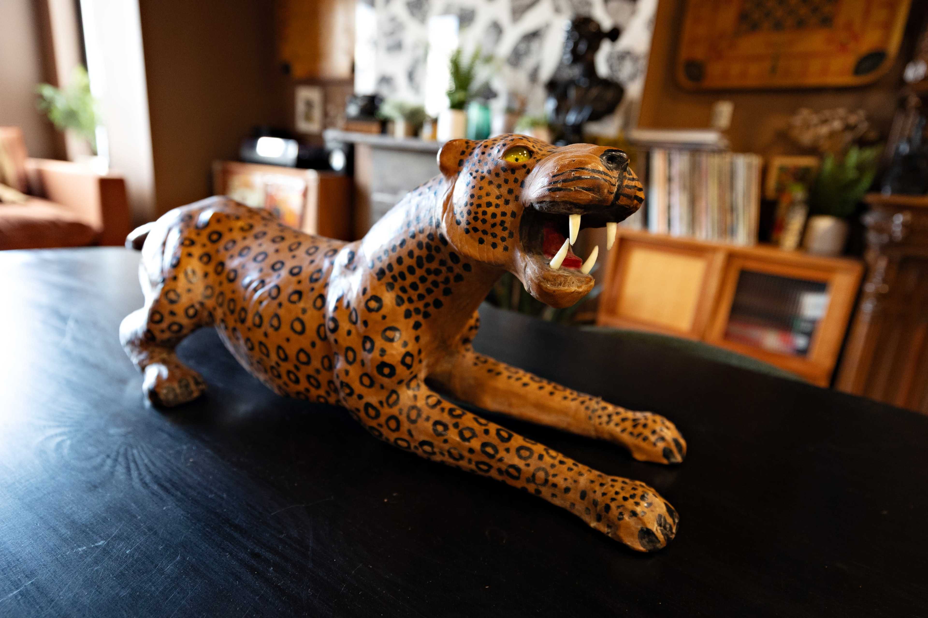 The image shows a decorative wooden sculpture of a leopard, positioned on a black table in an interior setting.