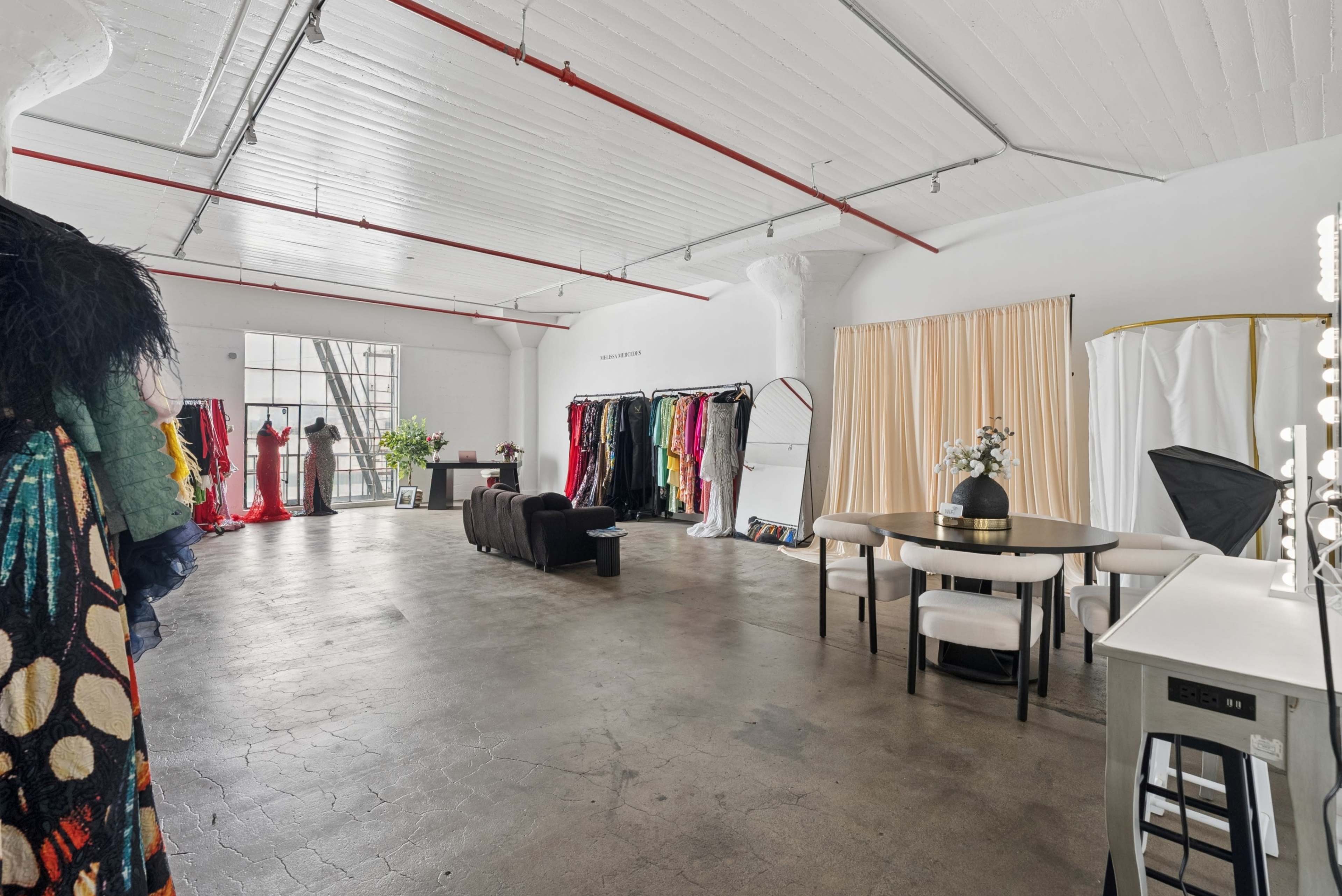 The image shows a spacious studio with various colorful outfits displayed on racks, a dining table with chairs, and large windows illuminating the area.