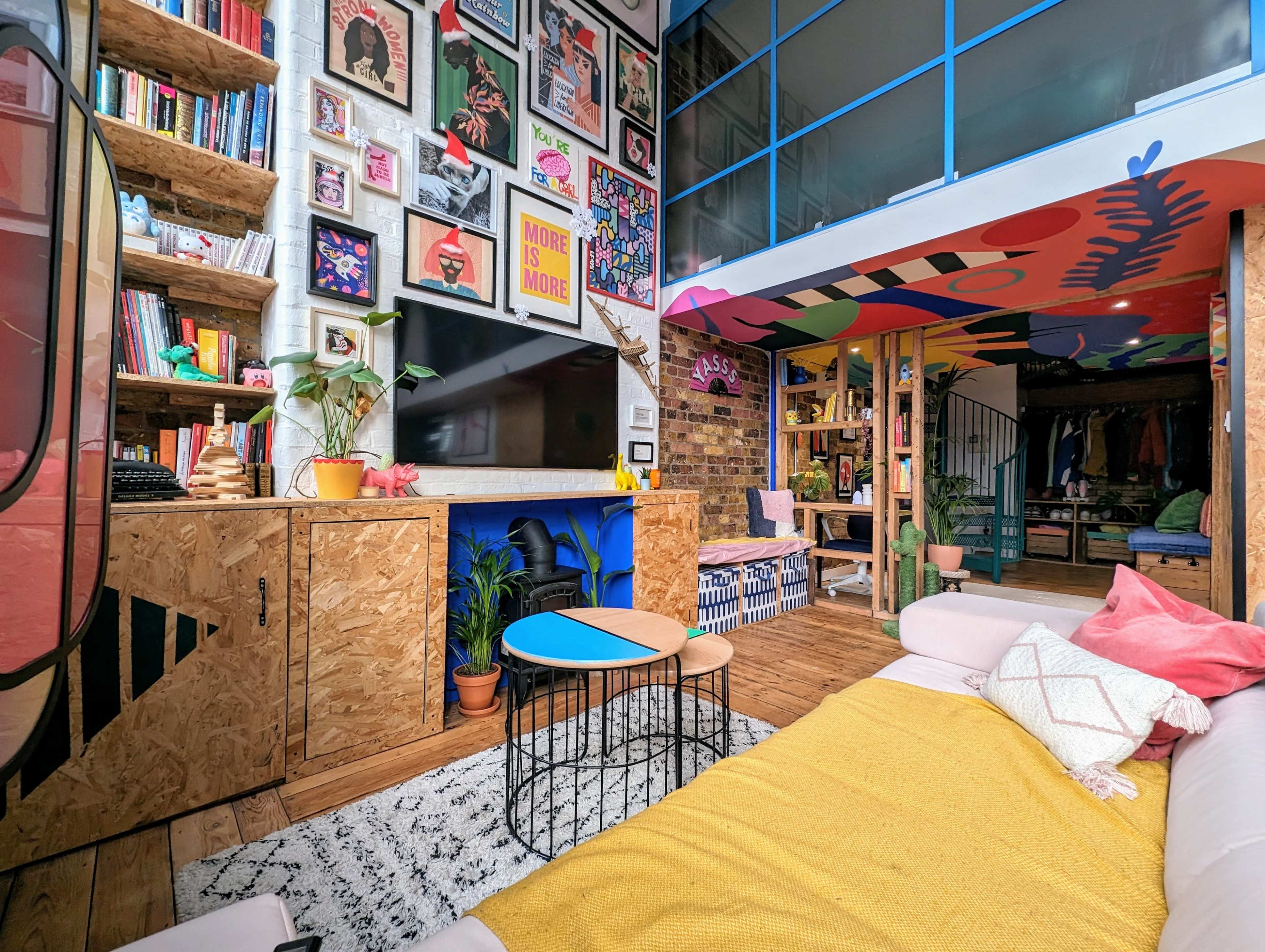 A colorful living room with a modern design, showcasing a wall adorned with framed artwork, a cozy seating area, and shelves filled with books and decorative items.