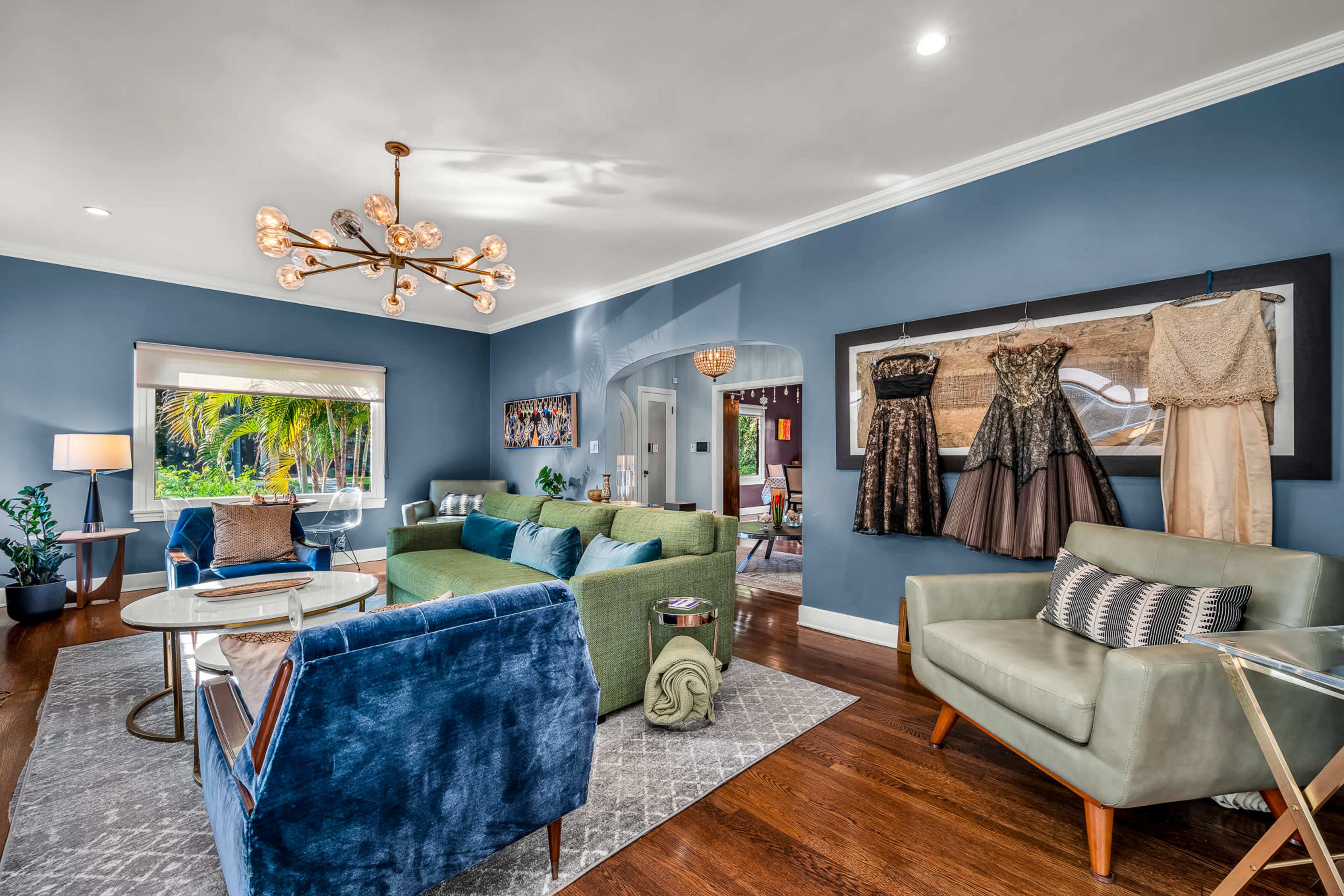 The image shows a stylish living room featuring an assortment of modern furniture, decorative dresses displayed on the wall, and large windows allowing natural light to enter.