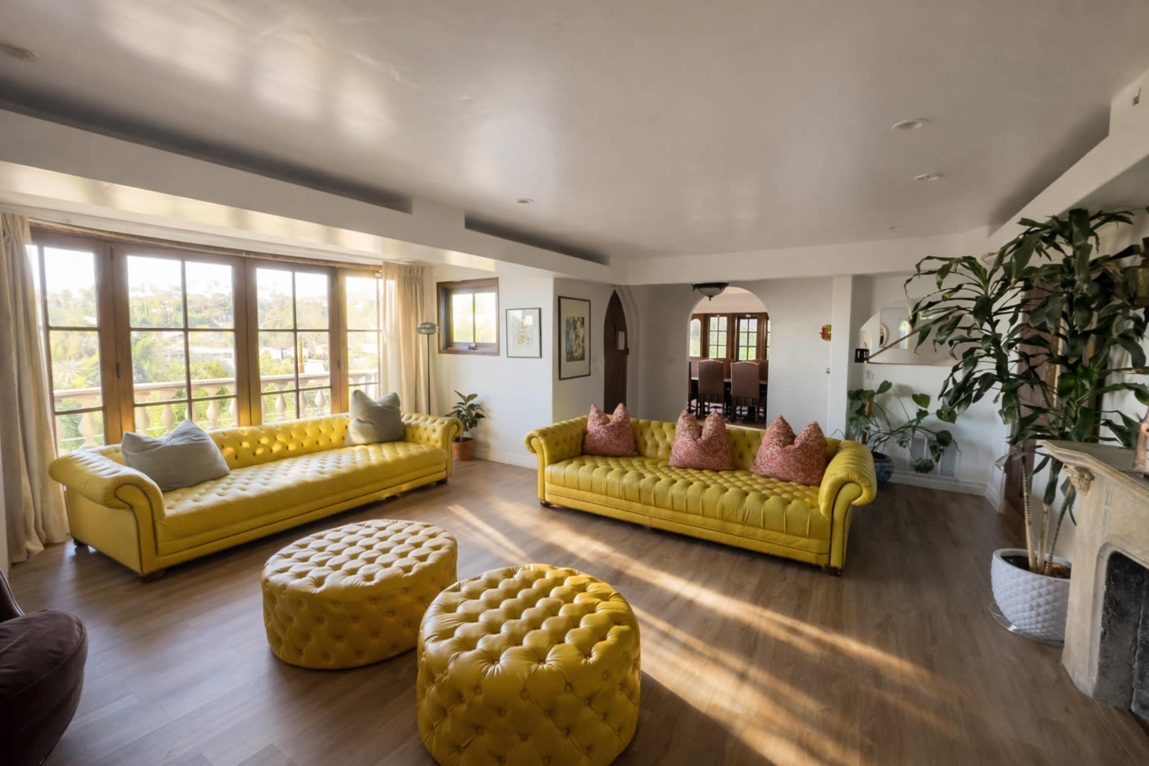 A spacious living room features two yellow upholstered sofas, round yellow ottomans, and large windows that allow natural light to fill the space.