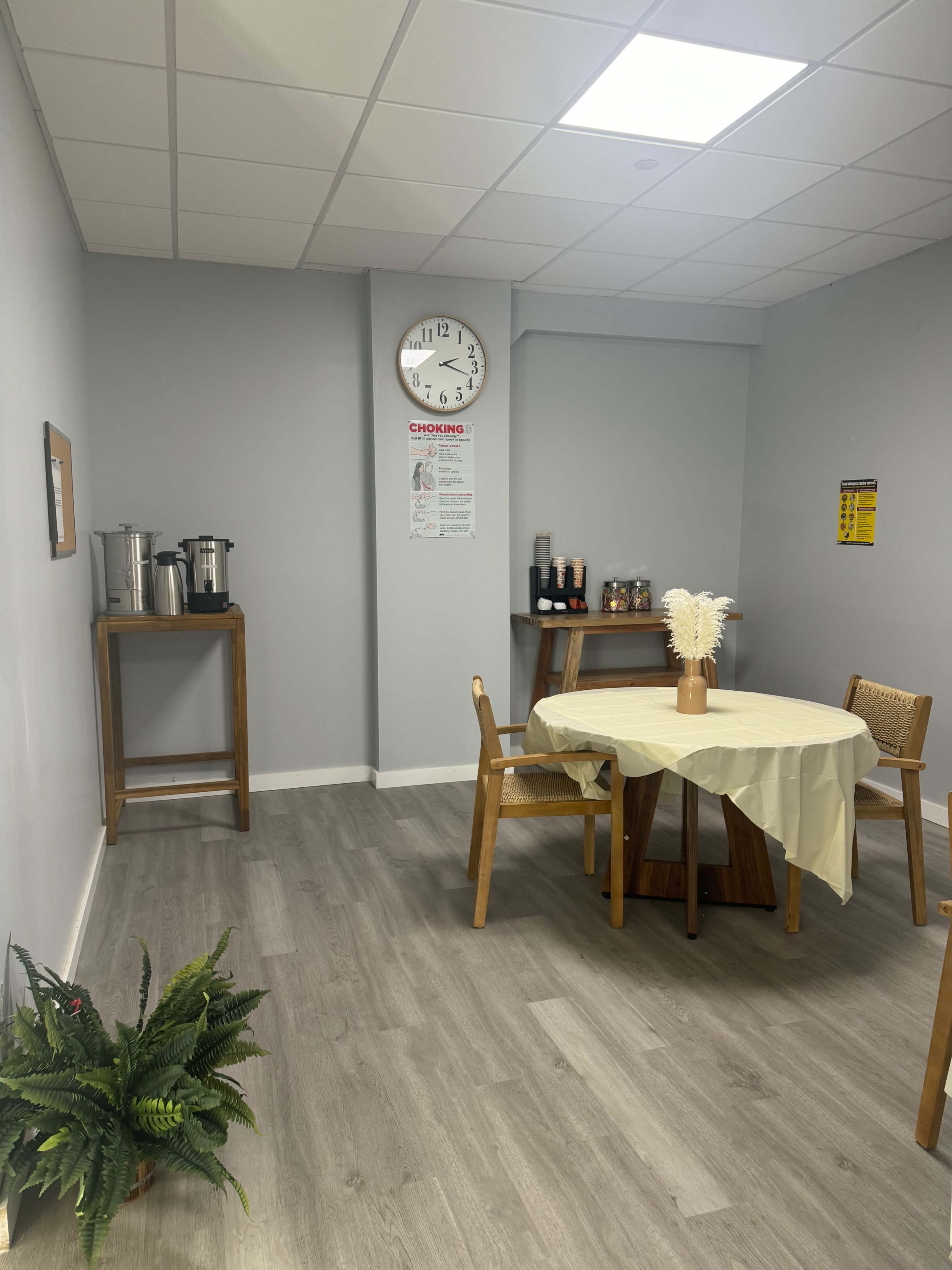 The image shows a simple break room with a round table, chairs, a clock on the wall, a small kitchenette with coffee dispensers, and a plant in the corner.