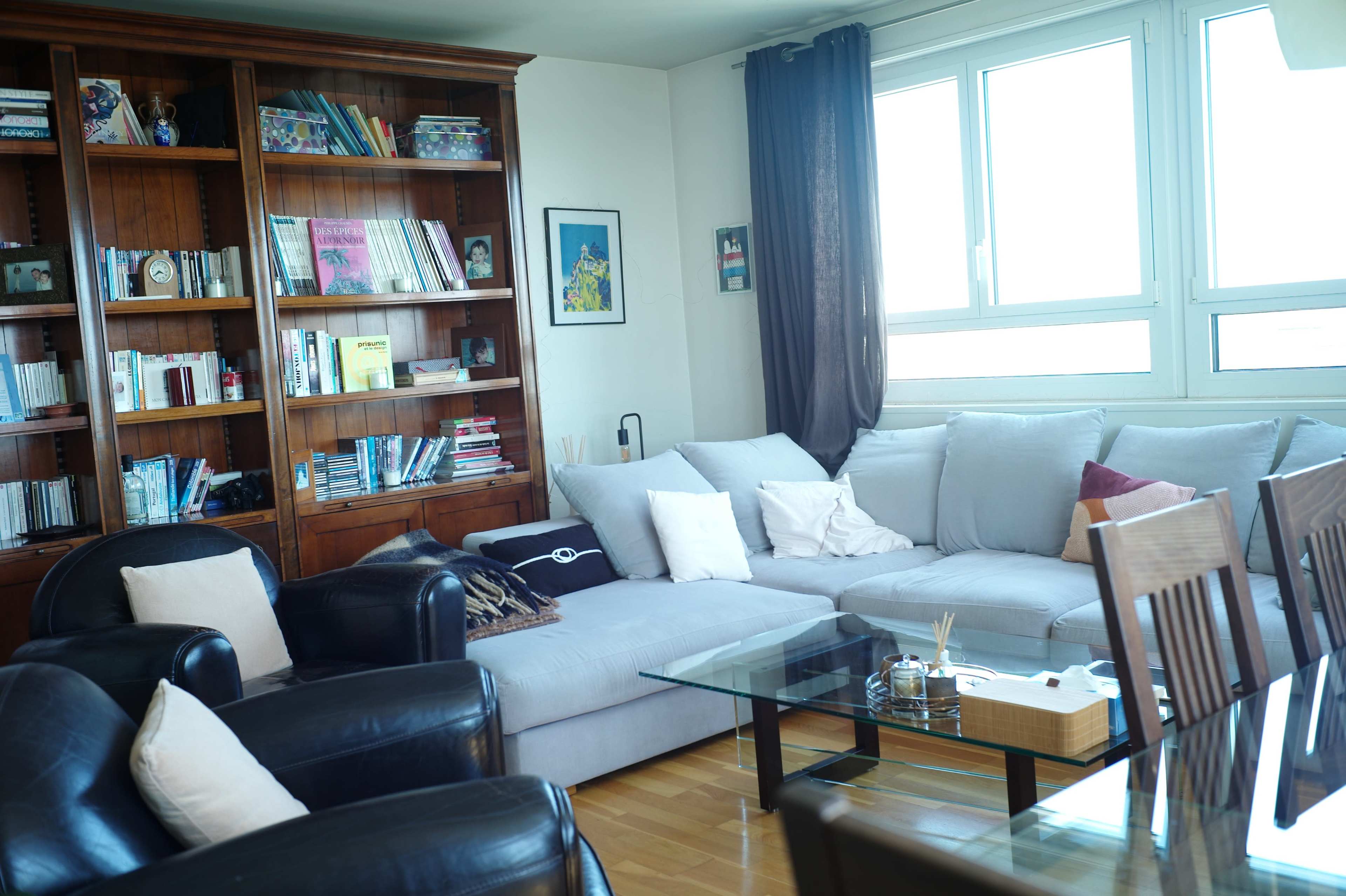 The image shows a living room with a gray sectional sofa, two black armchairs, a glass coffee table, and a wooden bookshelf filled with books and decorative items.