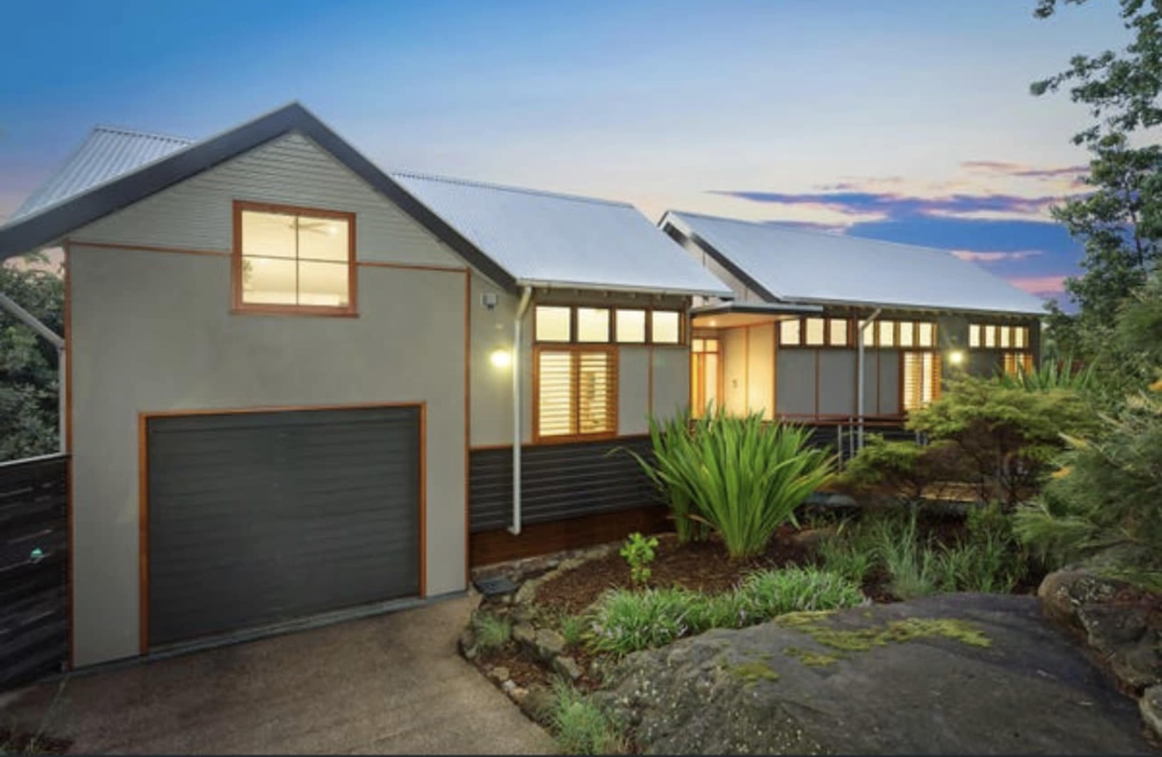 A modern house with a sloped roof and a garage sits amid landscaped greenery at dusk.